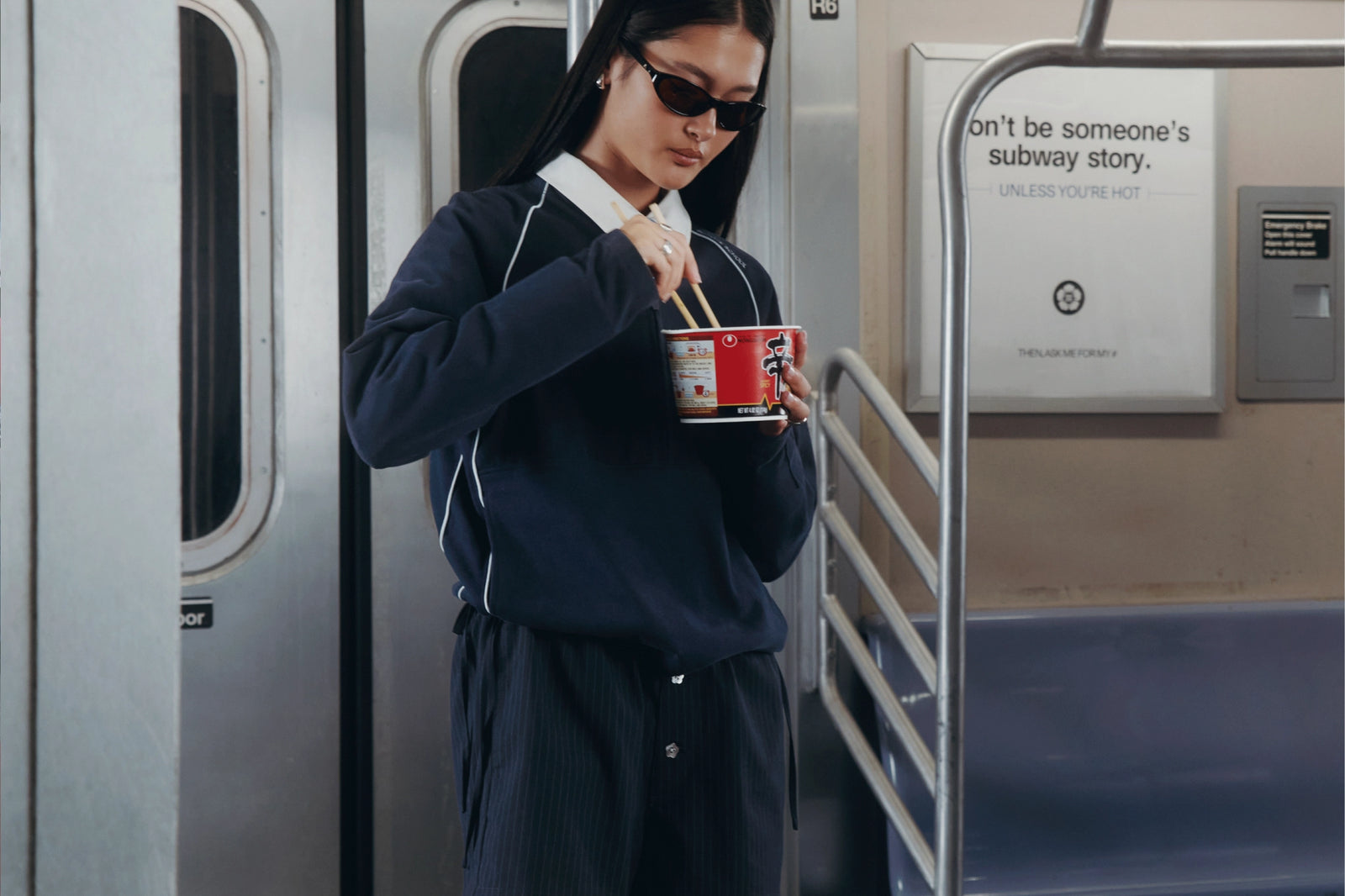 A woman wearing a dark navy blue tracksuit and black sunglasses stands on a subway train, holding a red cup with chopsticks. The background shows the metallic doors of the train and a sign with the text 'don't be someone's subway story.