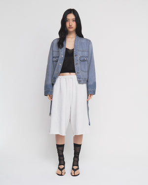 A woman stands against a white background wearing the MEDIUM BLUE TAEKWONDO DENIM JACKET by Sundae School, paired with a black top, loose white shorts with a detachable belt, and black strappy sandals with sheer black socks.