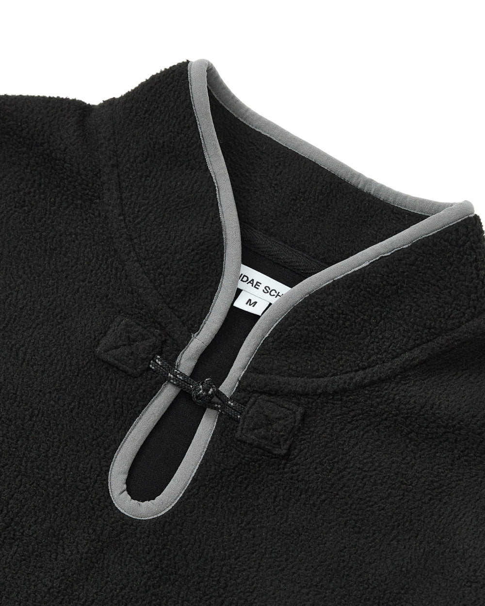 Close-up of the BLACK TEARDROP KNOT FLEECE by Sundae School, a black pullover inspired by East Asian design, featuring a mandarin collar with grey trim and a small looped button closure near the neck. A white Sundae School label showing size M is visible inside the collar.