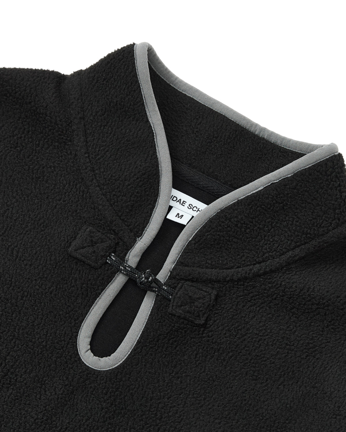 Close-up of the BLACK TEARDROP KNOT FLEECE by Sundae School, a black pullover inspired by East Asian design, featuring a mandarin collar with grey trim and a small looped button closure near the neck. A white Sundae School label showing size M is visible inside the collar.