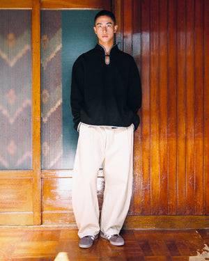 A person stands indoors against wood paneling, wearing the BLACK TEARDROP KNOT FLEECE by Sundae School, paired with wide beige pants and gray shoes. Their hands are in their pockets as they look at the camera.