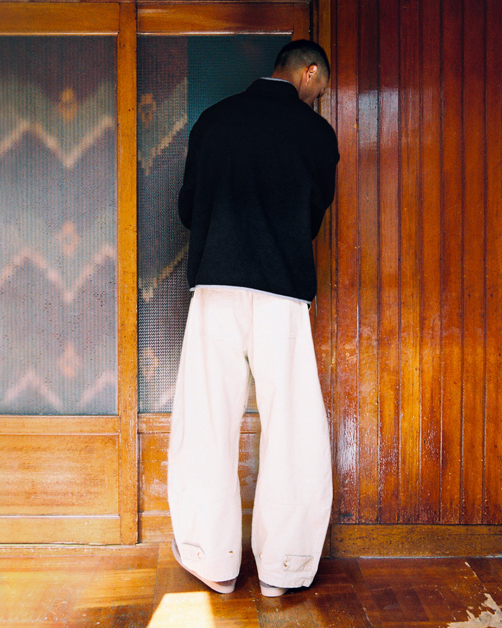 A person with short hair, dressed in a black jacket and wide beige pants, stands with their back to the camera before a wooden wall and door. Sunlight filters across the floor, softly showcasing their cozy BLACK TEARDROP KNOT FLEECE from Sundae School—a subtle nod to East Asian inspired fleece design.