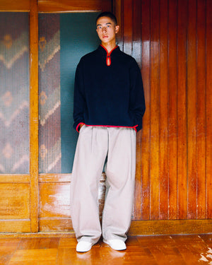 A person stands indoors against wooden sliding doors and patterned glass panels, wearing the Sundae School NAVY TEARDROP KNOT FLEECE—a fleece pullover with East Asian-inspired design details and red trim—paired with loose beige pants and white shoes.