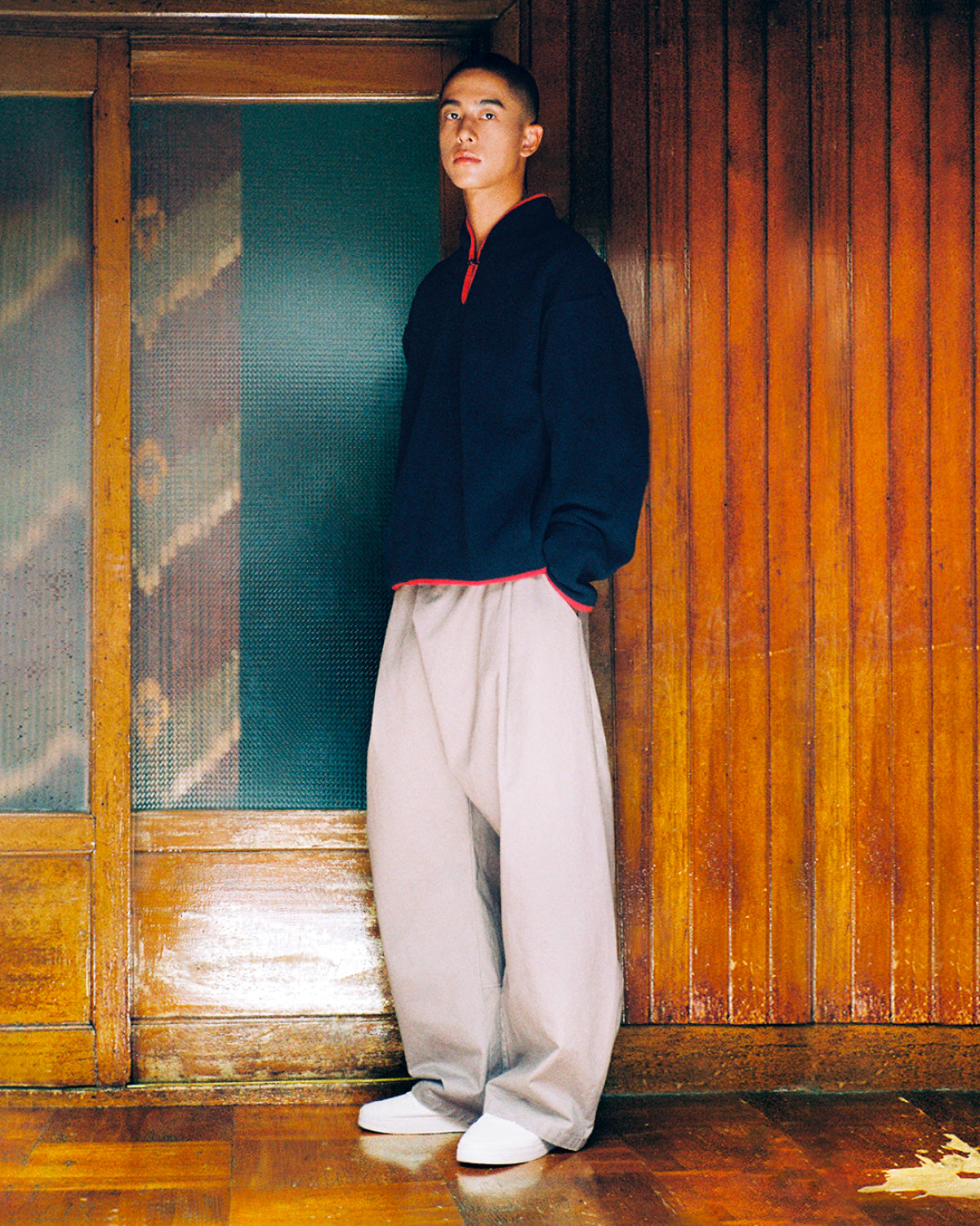 A person stands indoors on a wooden floor, wearing the Sundae School NAVY TEARDROP KNOT FLEECE over a red shirt, paired with loose light gray pants and white shoes, with a wooden wall and glass door in the background.