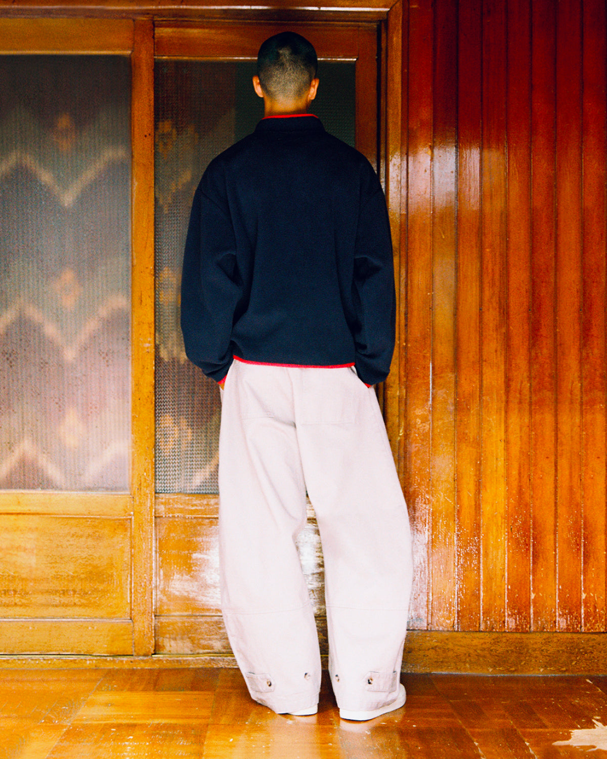 A person with short hair stands indoors, facing a wooden door, wearing the Sundae School NAVY TEARDROP KNOT FLEECE layered under a dark jacket and paired with loose, light-colored pants. The wooden walls and floor create a warm atmosphere that complements the cozy fleece details of their outfit.