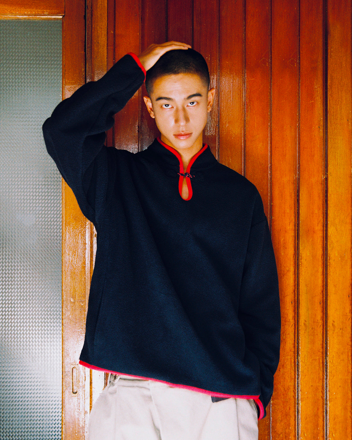 A person with short hair stands in front of a wooden wall, resting one hand on their head and looking at the camera while wearing the Sundae School NAVY TEARDROP KNOT FLEECE—an East Asian inspired long-sleeve top with red trim—paired with beige pants.