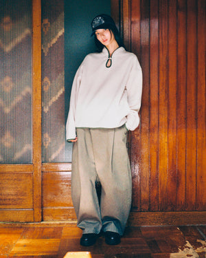 A person wearing Sundae School’s TAUPE TEARDROP KNOT FLEECE, paired with wide beige pants and a dark cap, stands indoors leaning casually against a wooden wall with a slight smile.