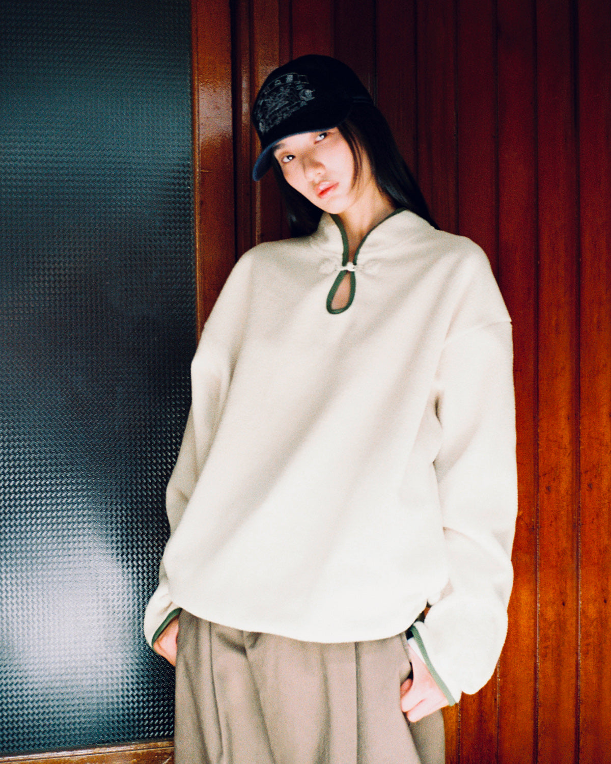 A person wearing the Sundae School TAUPE TEARDROP KNOT FLEECE, paired with brown wide-leg pants and a black cap, stands in front of a wooden wall, looking at the camera in a relaxed pose.