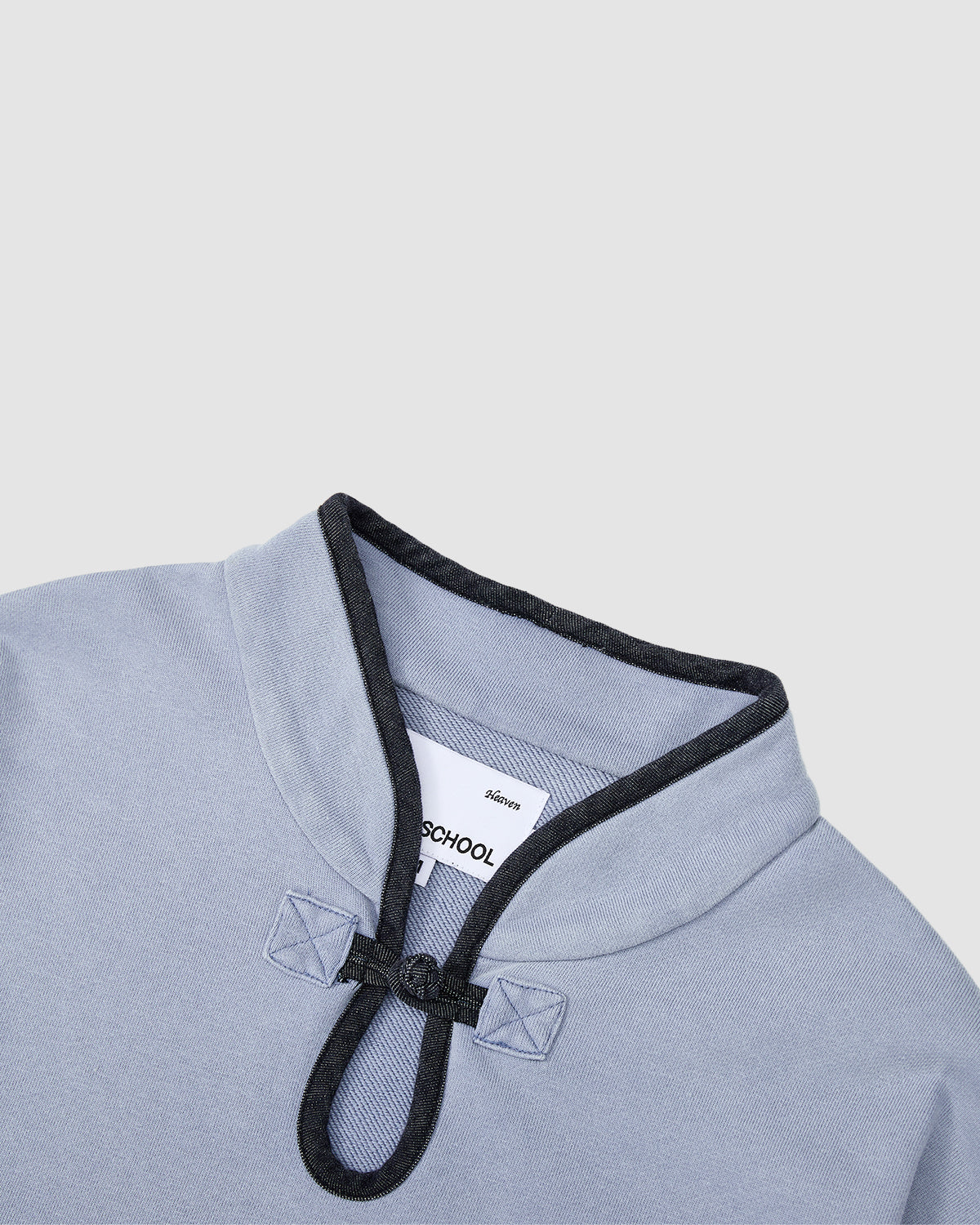 The SKY BLUE TEARDROP KNOT SWEATSHIRT by Sundae School is a light blue cotton sweatshirt with a distinctive collar featuring dark gray trim and a looped button closure. A white label is visible inside the collar against a plain, light gray background.
