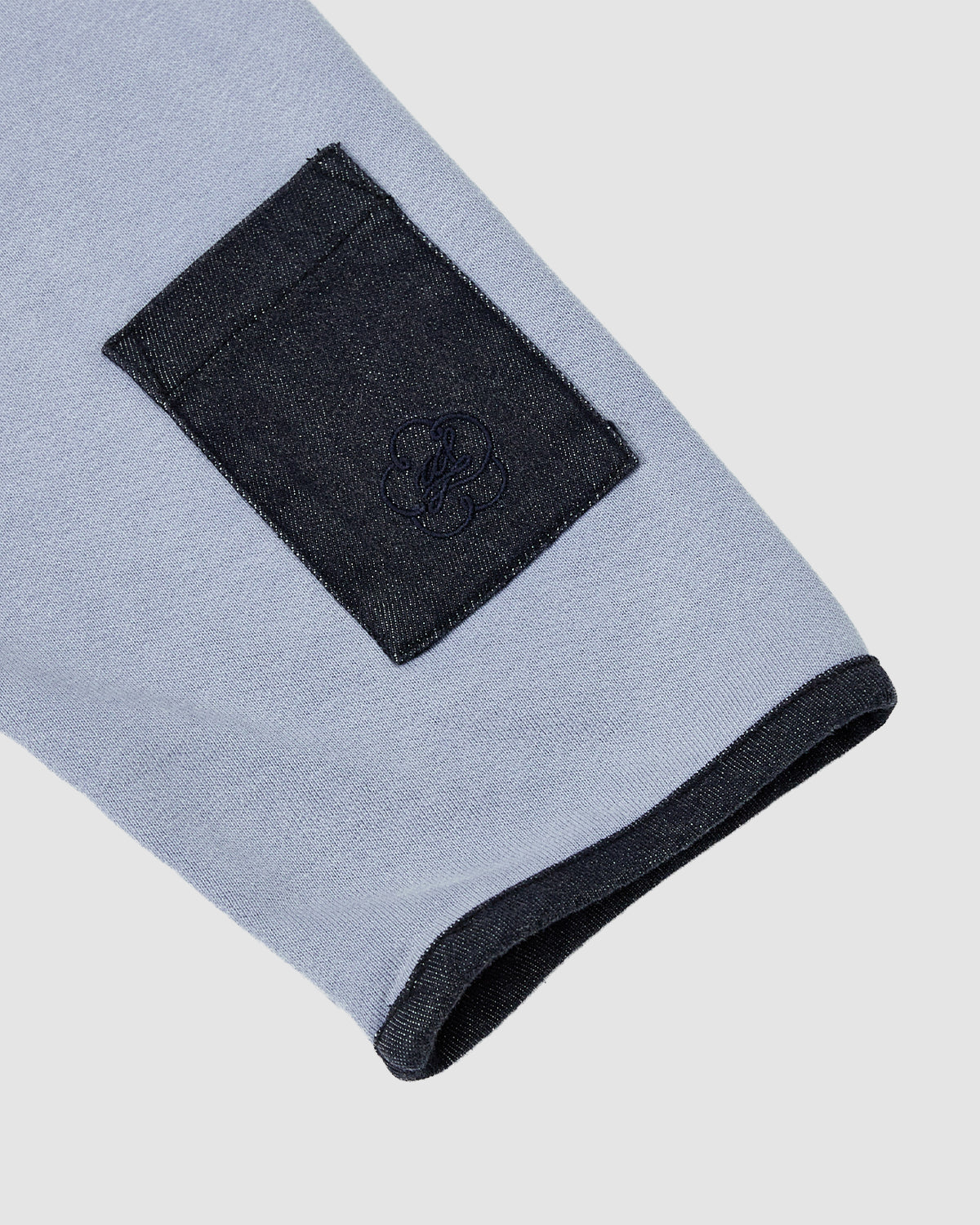 A close-up of the Sundae School SKY BLUE TEARDROP KNOT SWEATSHIRT sleeve, made from 100% cotton, showcases a dark denim trim patch and embroidered detail near the cuff, set against a plain light gray background.