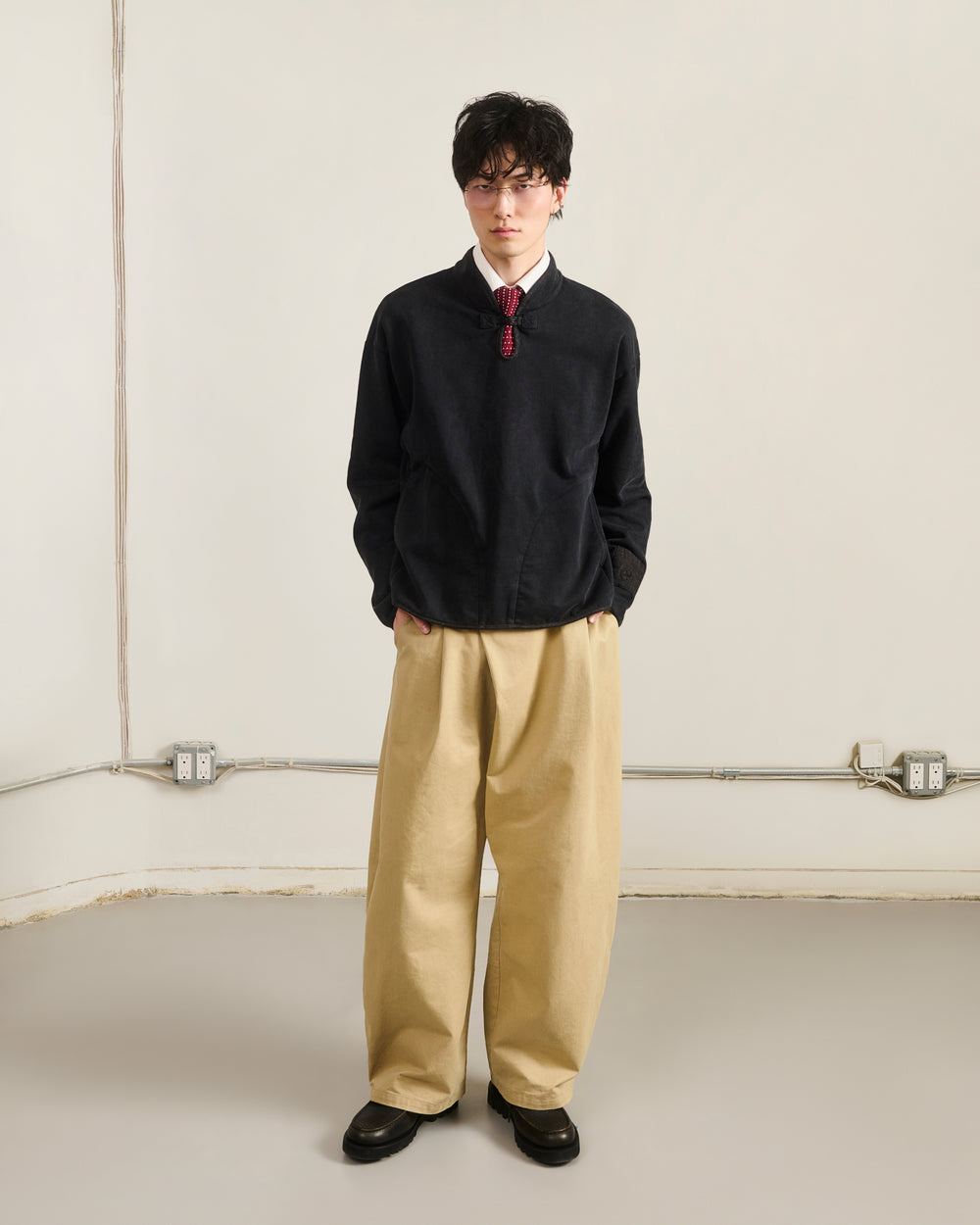 Indoors against a light wall, a person wears Sundae School’s BLACK TEARDROP KNOT SWEATSHIRT with beige wide-leg pants, black shoes, and glasses. Hands in pockets; electrical outlets and cables are visible on the wall.