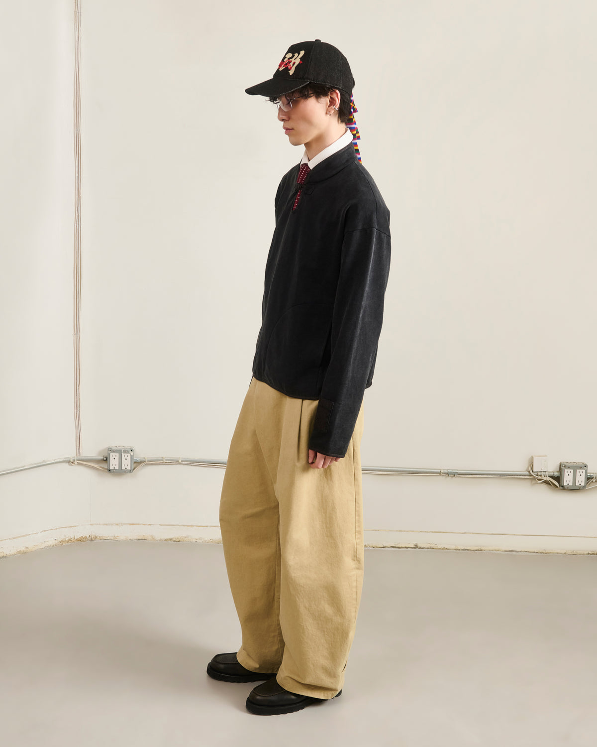 A person stands in a minimalist room wearing braided hair, a black cap, glasses, wide beige pants, black shoes, and the Sundae School BLACK TEARDROP KNOT SWEATSHIRT over a white collared shirt with a red tie, facing sideways.