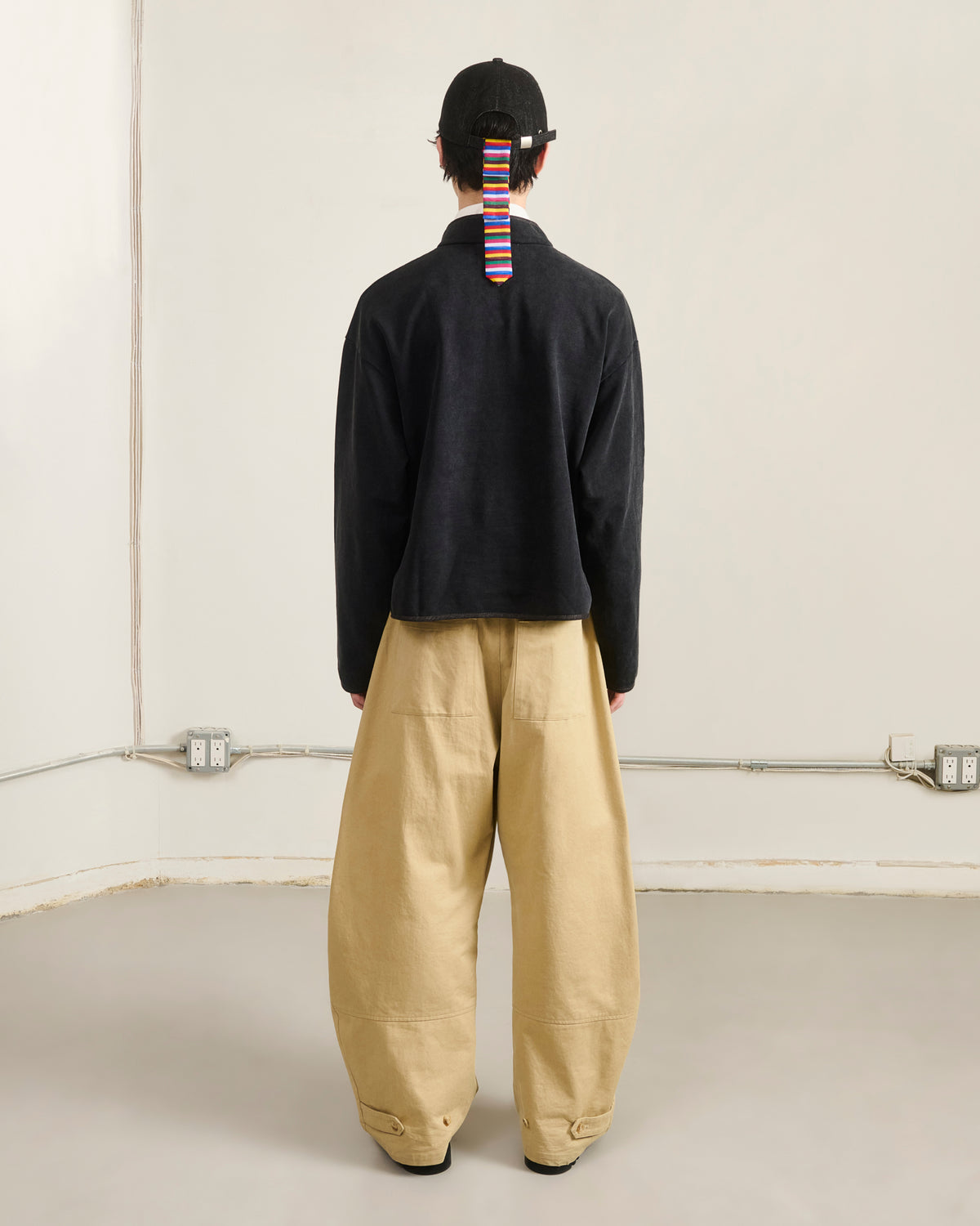 A person stands facing away in an empty, minimal indoor room wearing Sundae School’s BLACK TEARDROP KNOT SWEATSHIRT beneath a black jacket, with wide beige pants, black shoes, and a black cap featuring a colorful striped ribbon.