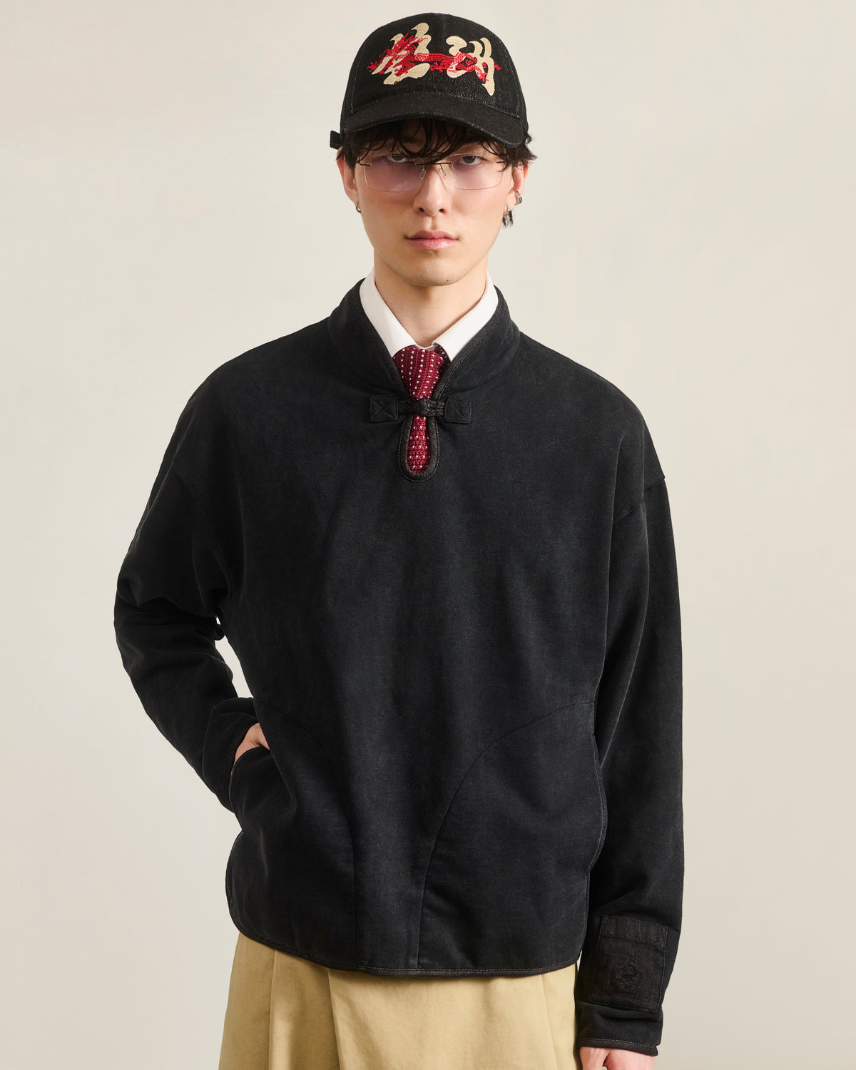 A person stands against a plain light background wearing Sundae School’s BLACK TEARDROP KNOT SWEATSHIRT layered under a black jacket, styled with a black baseball cap, glasses, white shirt, red-patterned tie, and beige pants.