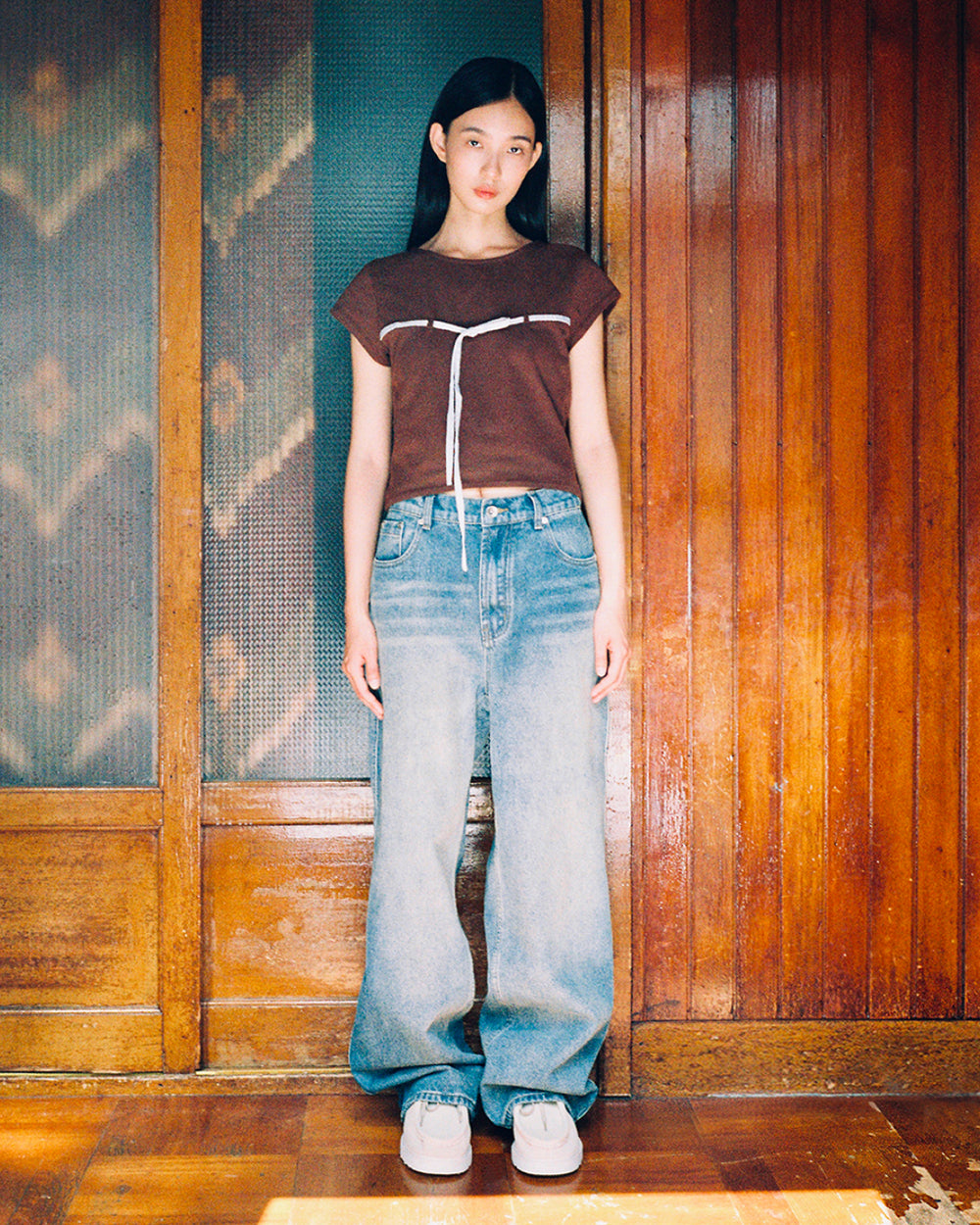 A woman stands indoors wearing the Sundae School BROWN TIE GORUM BABY TEE, a brown cropped baby tee with a scooped neckline, paired with oversized blue jeans and white sneakers. She poses against wooden paneled doors, looking at the camera with a neutral expression.