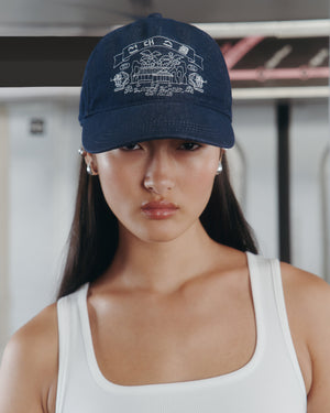 A young woman with long dark hair wears the Sundae School INDIGO DENIM TIGER CAP, a navy adjustable six-panel cap with white graphic designs, paired with a white tank top and standing indoors with a neutral expression.