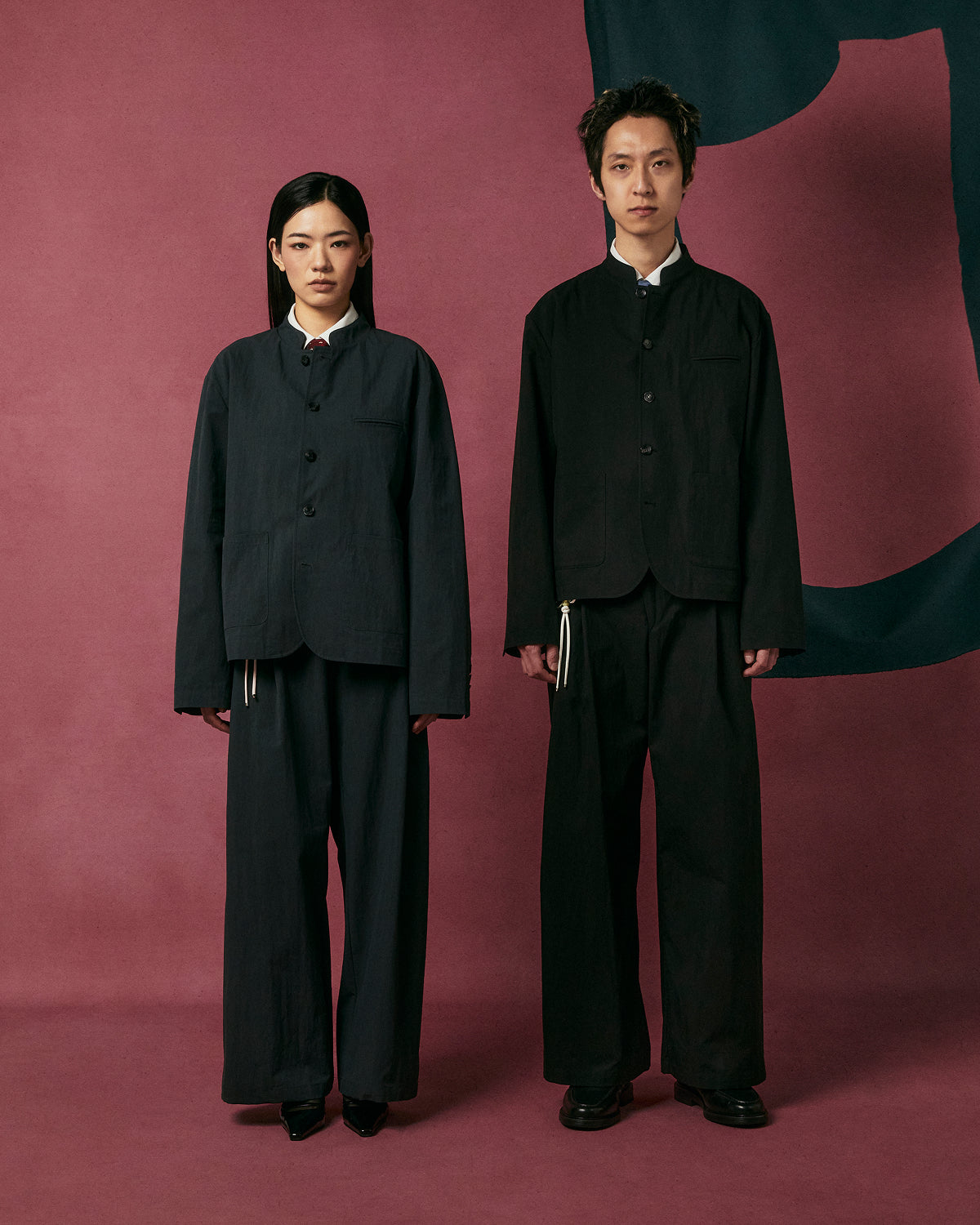 Two people stand side by side against a burgundy backdrop, both wearing Sundae School's CHARCOAL PLEATED UNIFORM SLACKS, looking directly at the camera with neutral expressions.