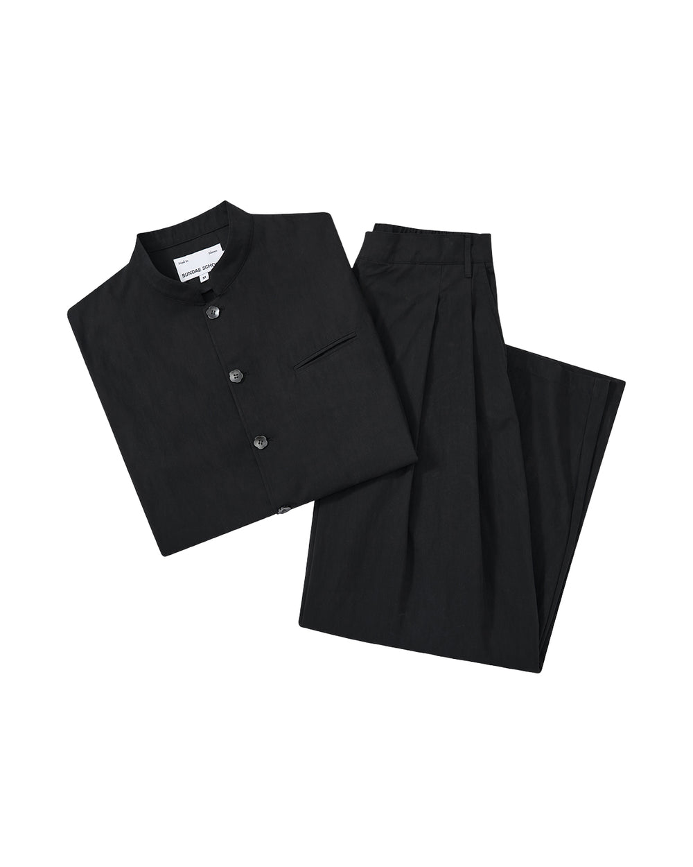 The BLACK PLEATED UNIFORM SLACKS by Sundae School are shown neatly folded with a black mandarin-collar shirt, both displayed on a plain light gray background.