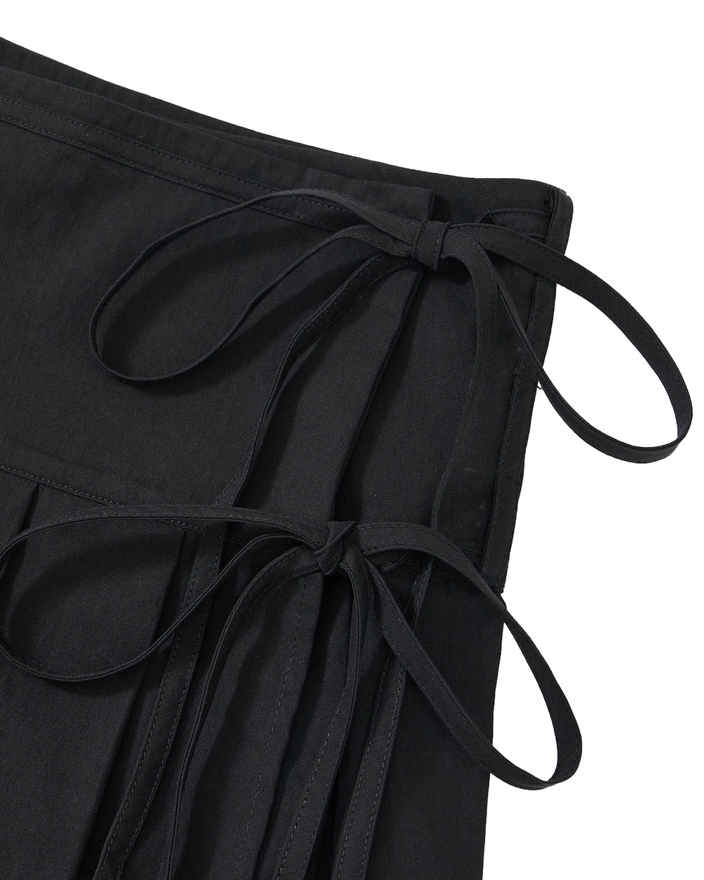 Close-up of the Sundae School BLACK UNIFORM APRON SKIRT, a black pleated skirt inspired by Korean-style aprons. It features two fabric ties on the waistband, neatly knotted into bows, and is crafted from cotton or a similar material with visible stitching along the edges.