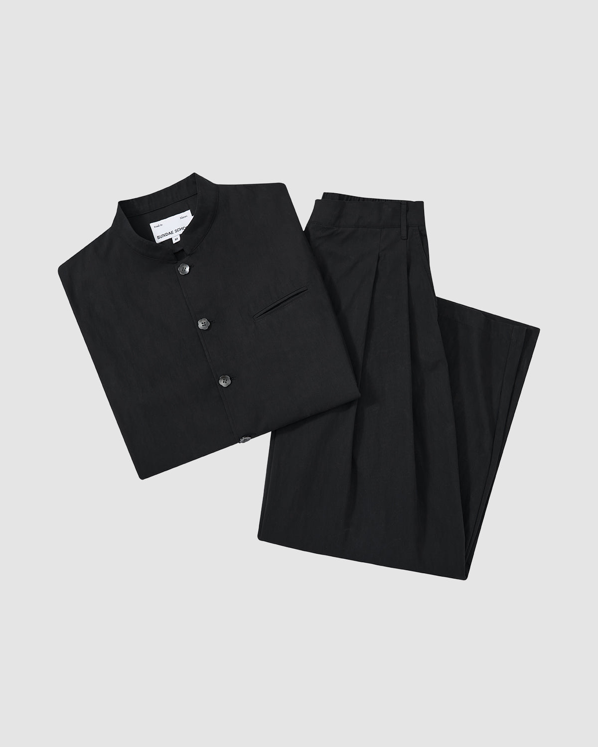 The BLACK PLEATED UNIFORM SLACKS by Sundae School are shown neatly folded with a black mandarin-collar shirt, both displayed on a plain light gray background.