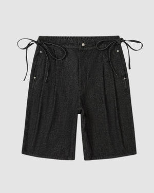 The BLACK VITA JORTS by Sundae School are black wide-leg denim shorts with an elastic waistband, side pleats, adjustable ties, and two metal snap buttons on each side at the waist. Shown against a plain light grey background.