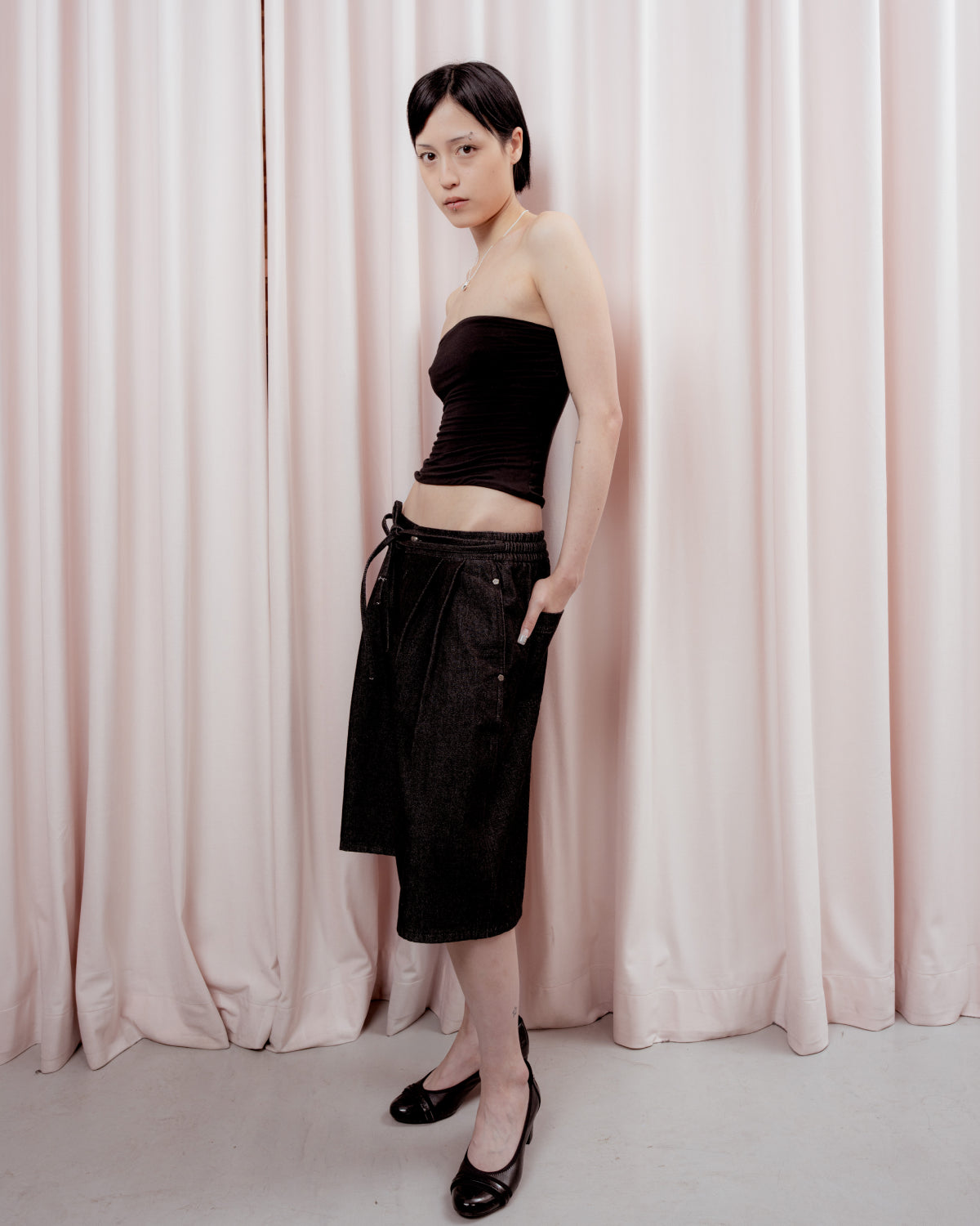 A person stands in front of pale pink curtains, wearing a black tube top, Sundae School’s BLACK VITA JORTS—wide-leg silhouette pleated denim jorts that fall to the knees—and black heels, with hands in pockets and head slightly tilted.