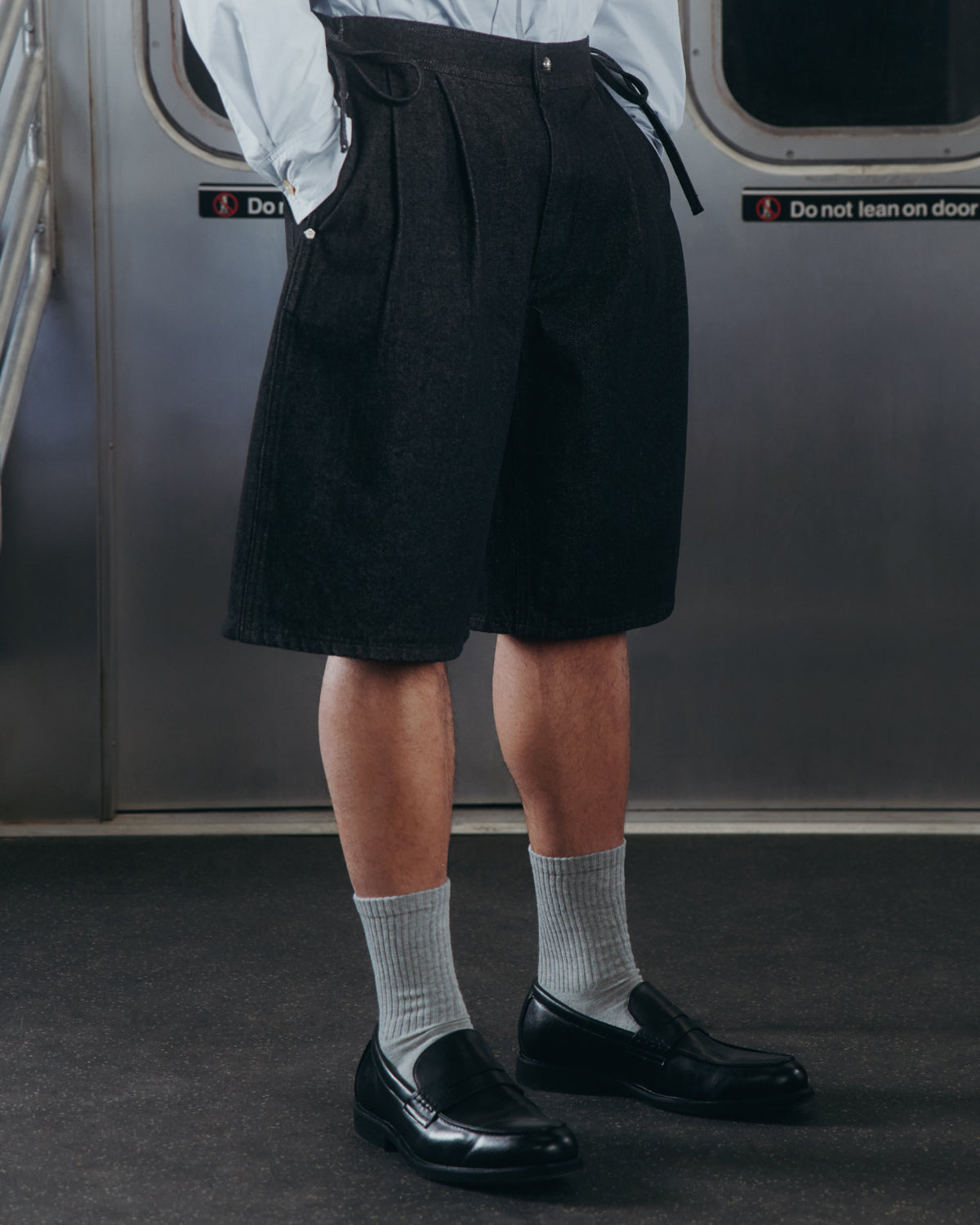 A person stands in a subway car wearing BLACK VITA JORTS by Sundae School, paired with light gray socks, black loafers, and a light blue shirt. Behind them is a subway door marked with caution signs.
