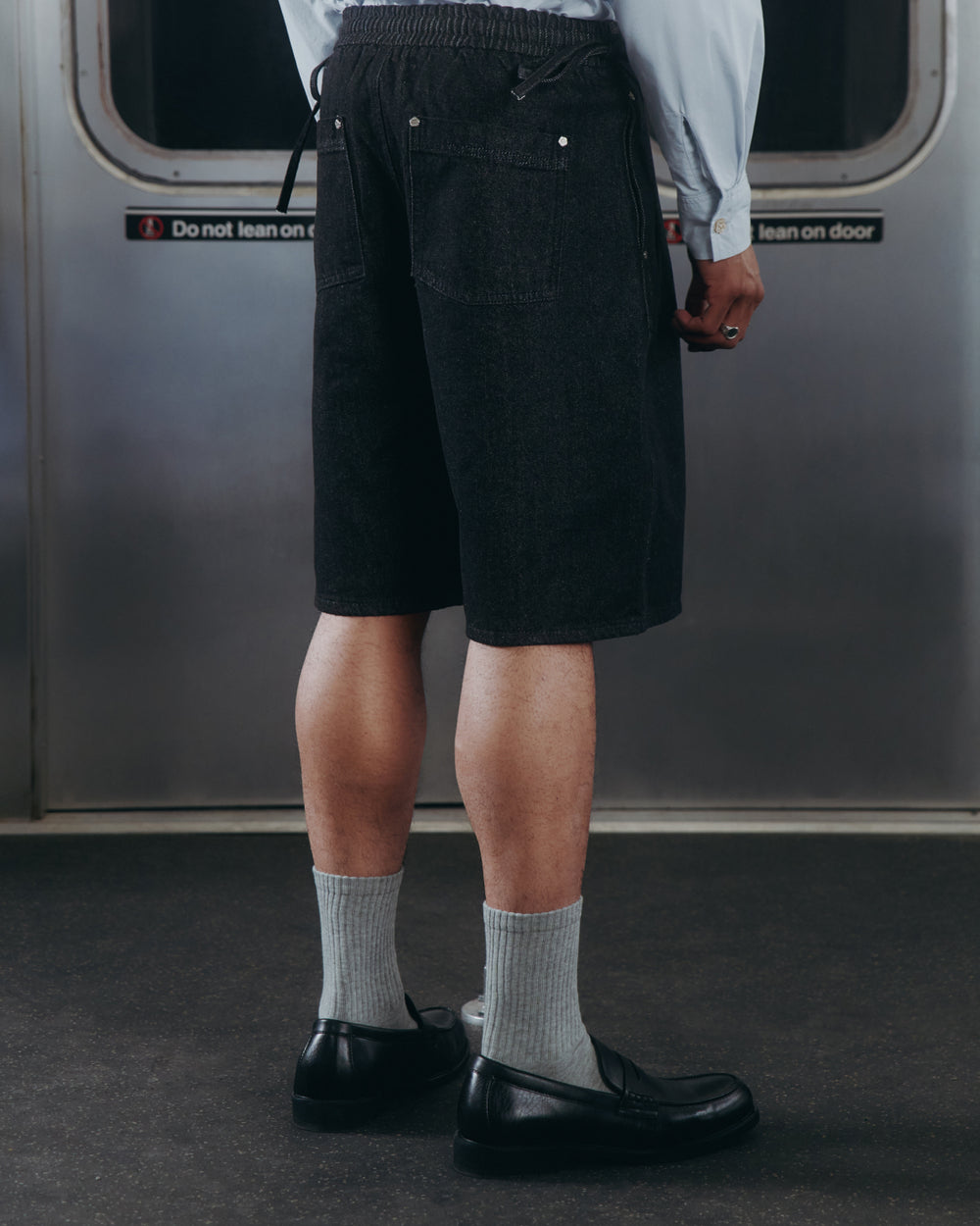 A person in a light blue shirt, Sundae School BLACK VITA JORTS, gray socks, and black shoes stands facing away on a subway train with a "Do not lean on door" sign visible in the background.