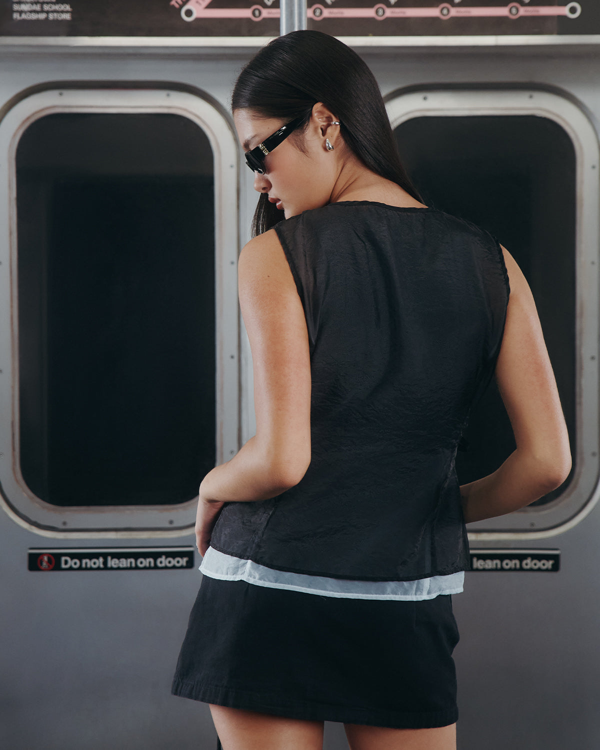A woman with long dark hair, wearing sunglasses and a Sundae School BLACK VITA ORGANZA VEST over a black skirt, stands facing away inside a subway train near doors labeled 