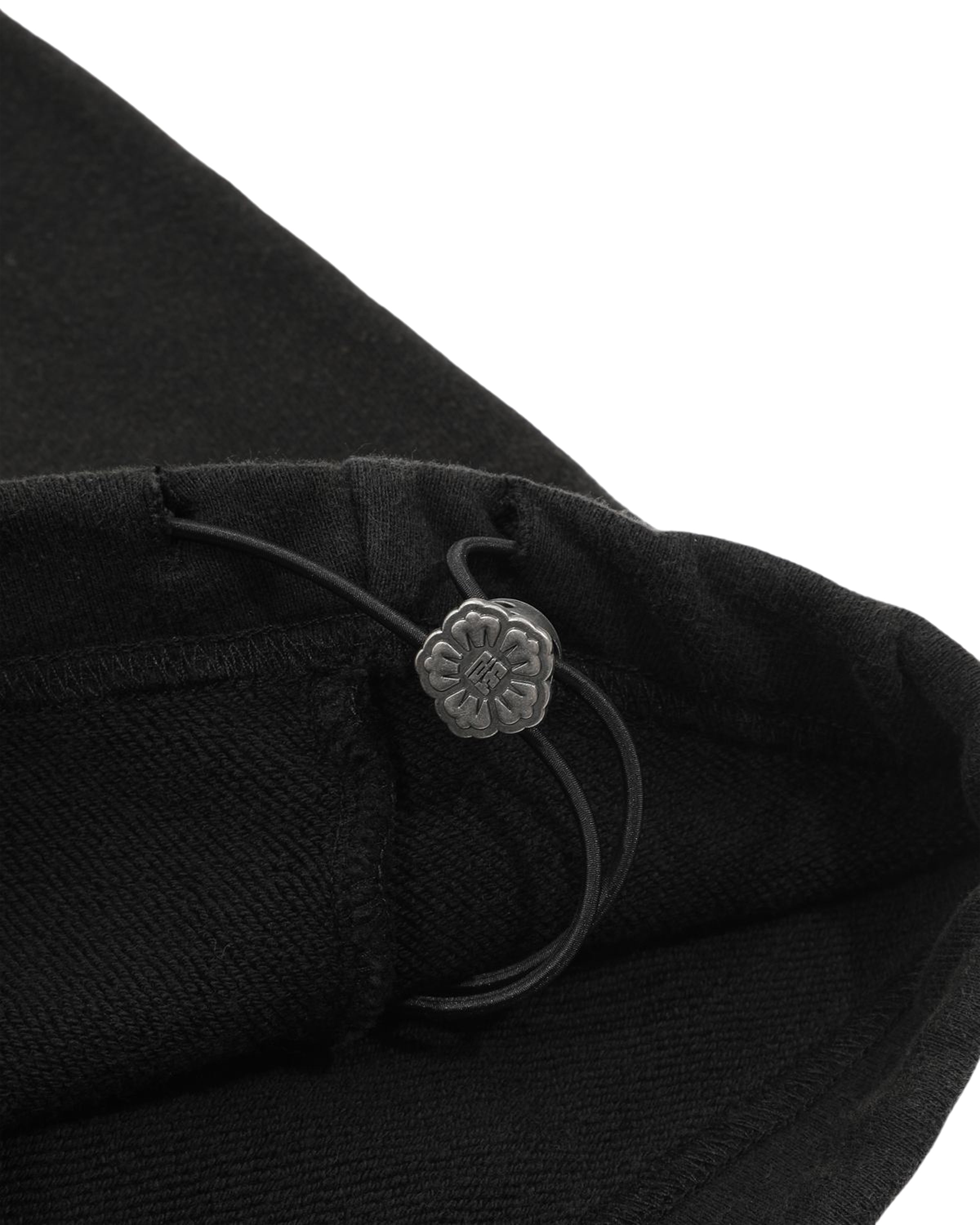 Close-up of the black fabric from Sundae School's BLACK WASH WRAP SWEAT BAJI, featuring a small, flower-shaped metal button attached to an elastic cord—likely part of the adjustable wrap-front drape detail.