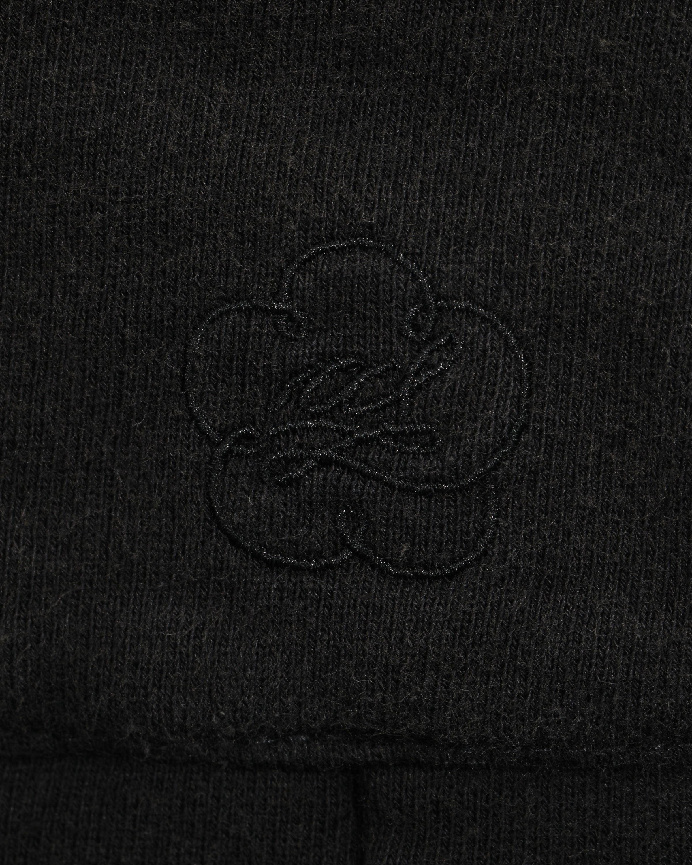 Close-up of the black fabric from the Sundae School BLACK WASH WRAP SWEAT BAJI, featuring a subtle, stitched clover-shaped logo design in the center. The texture of the material and the embroidery are clearly visible, highlighting the quality craftsmanship typical of these premium cotton sweatpants.