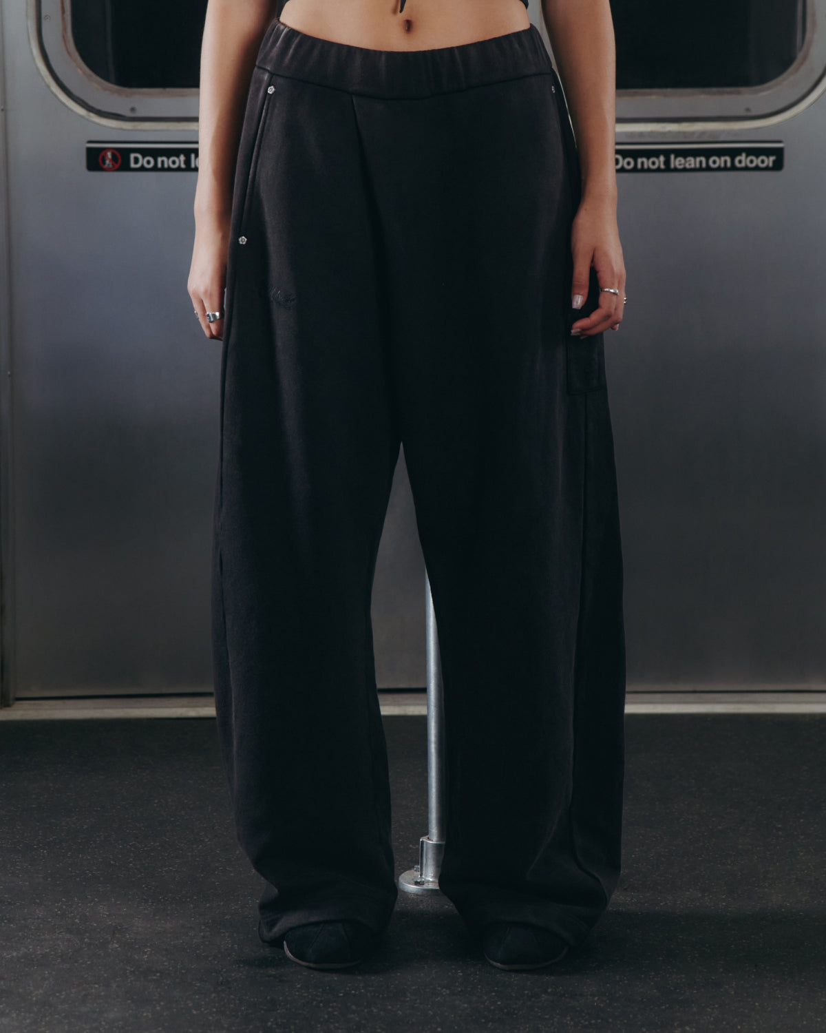 A person wearing Sundae School’s BLACK WASH WRAP SWEAT BAJI and black shoes stands inside a subway car with their hands at their sides. The upper body is not visible, and signs on the subway door behind read “Do not block” and “Do not lean on door.”.