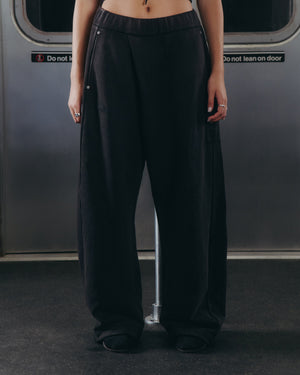 A person wearing Sundae School’s BLACK WASH WRAP SWEAT BAJI and black shoes stands inside a subway car with their hands at their sides. The upper body is not visible, and signs on the subway door behind read “Do not block” and “Do not lean on door.”.
