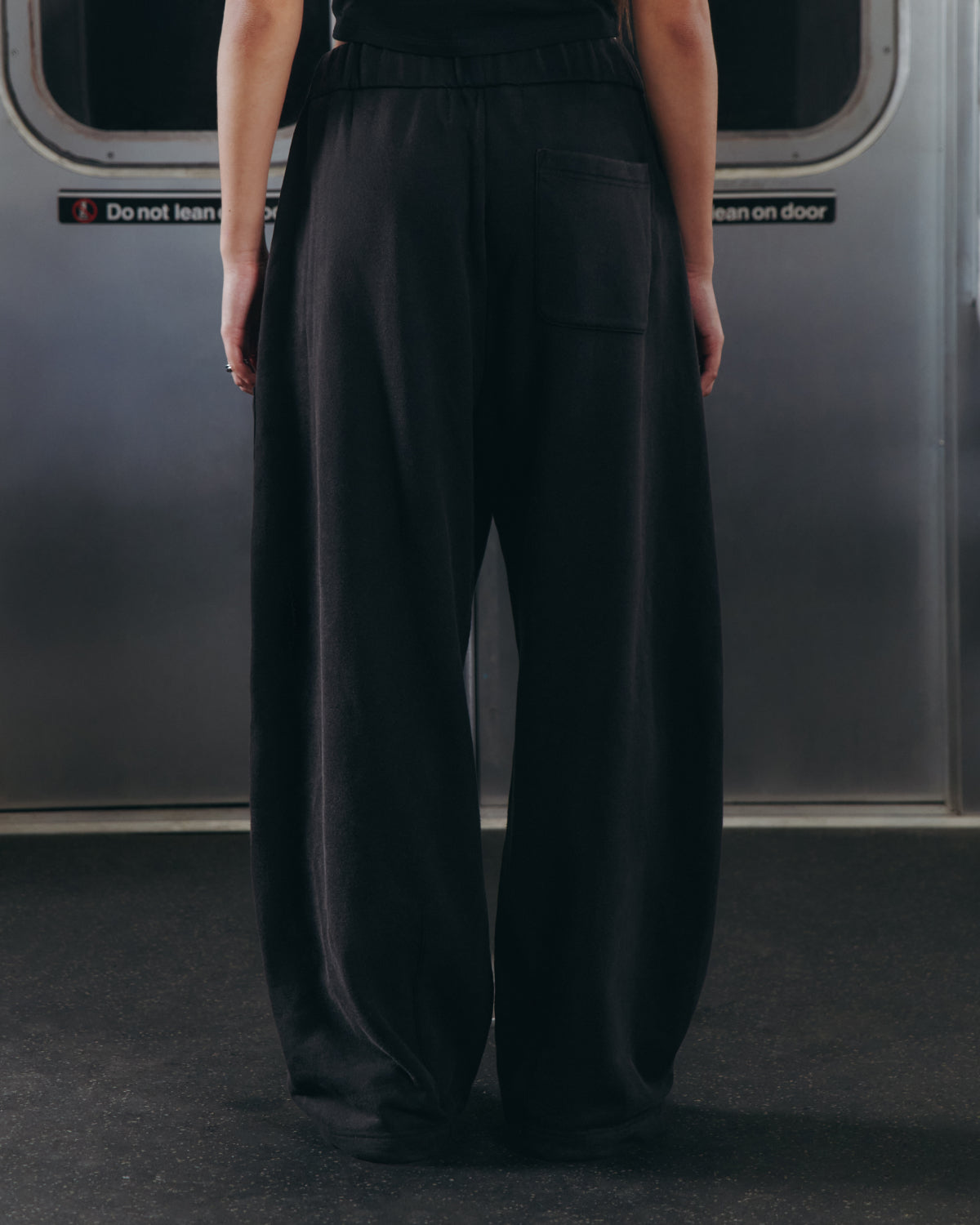A person wearing Sundae School’s BLACK WASH WRAP SWEAT BAJI stands with their back to the camera on a subway train, facing closed metal doors adorned with warning signs.