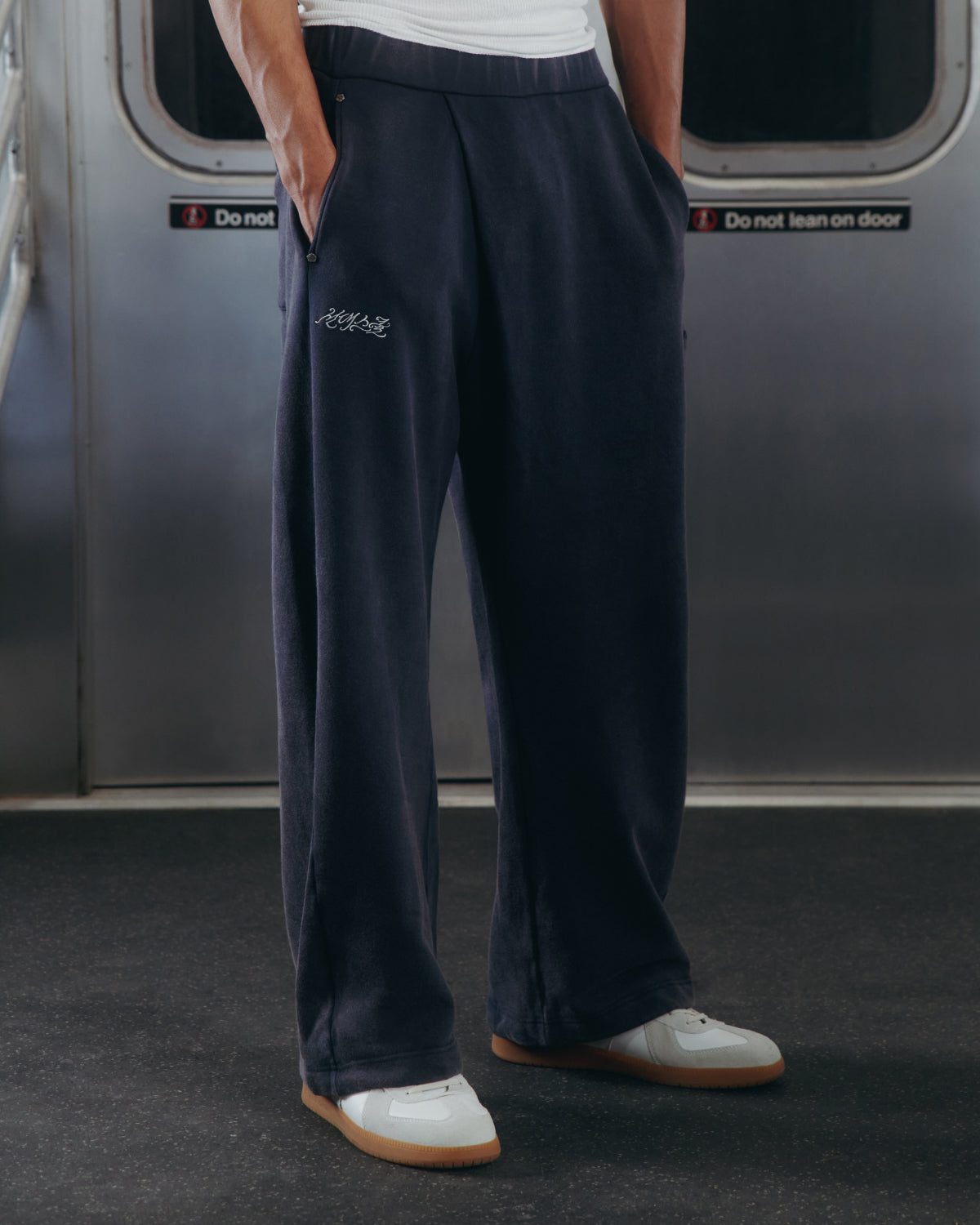 A person wearing Sundae School's NAVY WASH WRAP SWEAT BAJI with white sneakers stands on a subway train, hands in pockets, as 