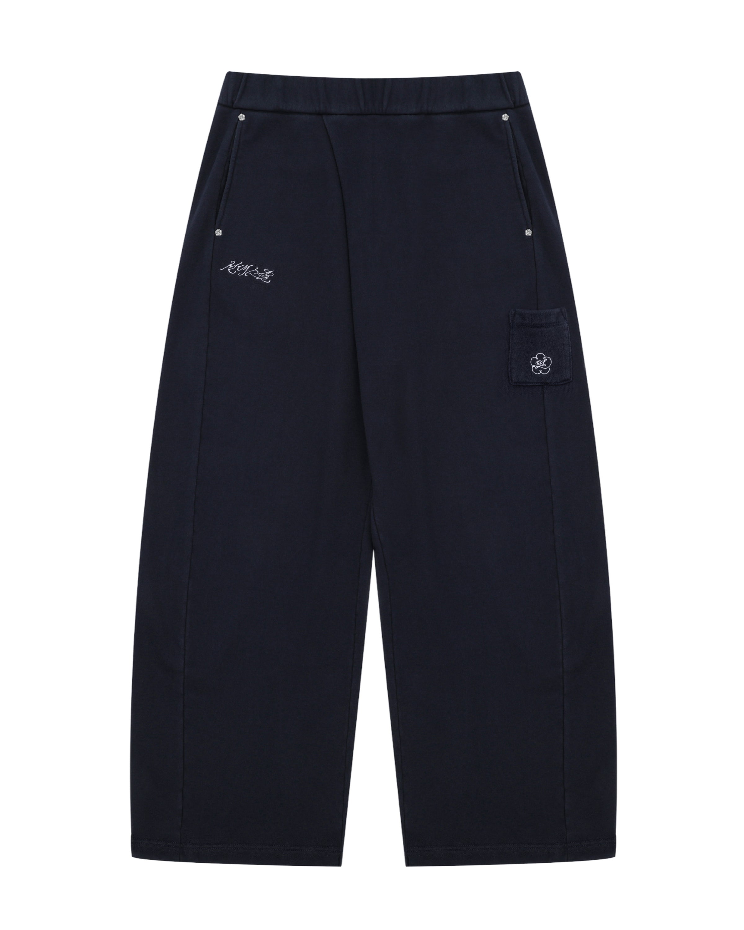 NAVY WASH WRAP SWEAT BAJI by Sundae School features a wide-leg unisex silhouette in black, an elastic waistband, two front pockets with button closures, subtle white embroidered text on one side, and a flap pocket adorned with a flower patch on the other.