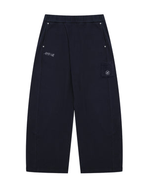NAVY WASH WRAP SWEAT BAJI by Sundae School features a wide-leg unisex silhouette in black, an elastic waistband, two front pockets with button closures, subtle white embroidered text on one side, and a flap pocket adorned with a flower patch on the other.