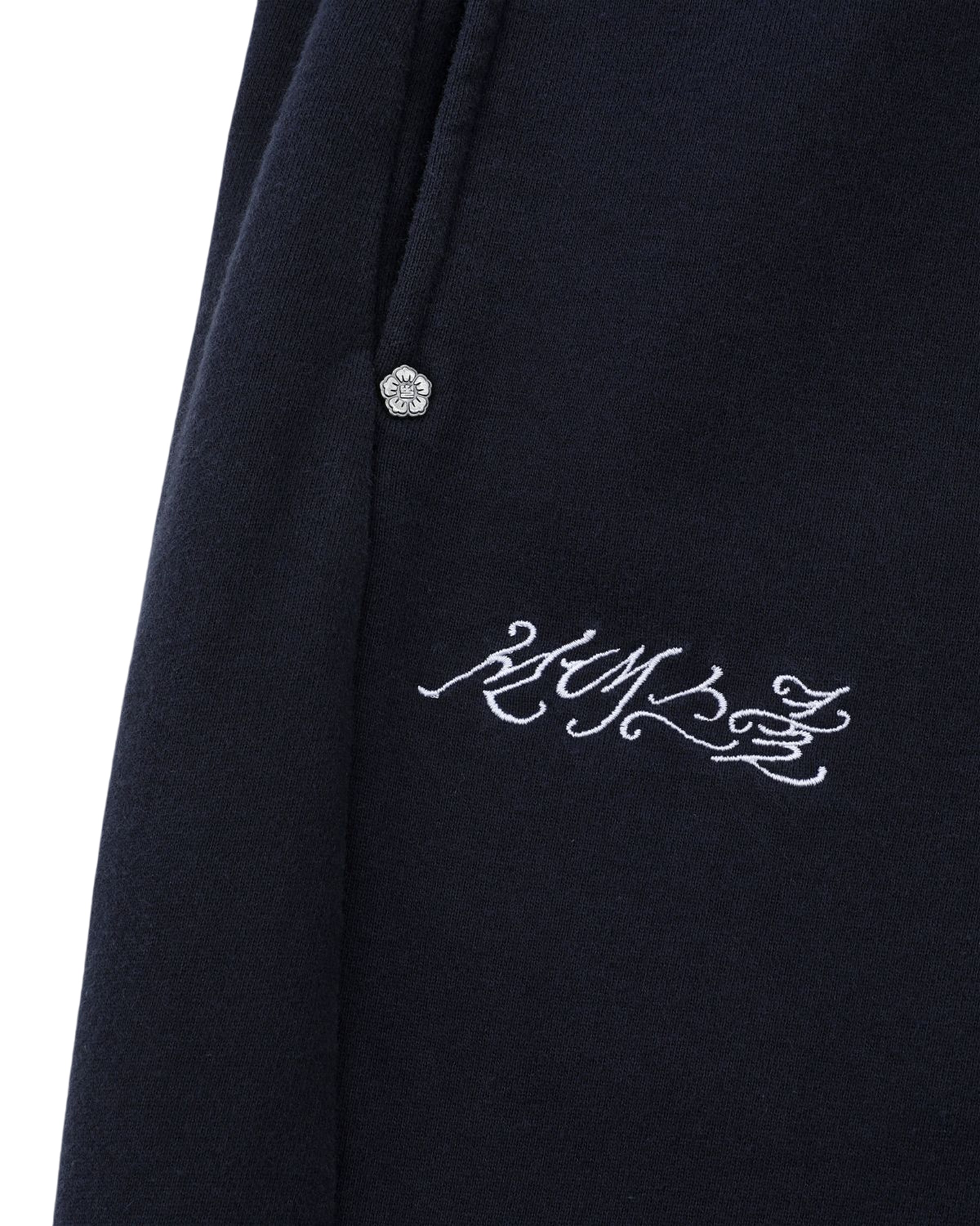 Close-up of Sundae School's NAVY WASH WRAP SWEAT BAJI––navy cotton sweatpants featuring a silver decorative button near the pocket and ornate white embroidery reading 