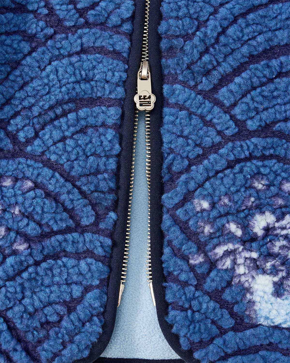 Close-up of a silver zipper on the Sundae School BLUE WAVE FLEECE, showing embossed pile with a blue and purple Korean ocean pattern, partially unzipped to reveal light blue lining underneath.