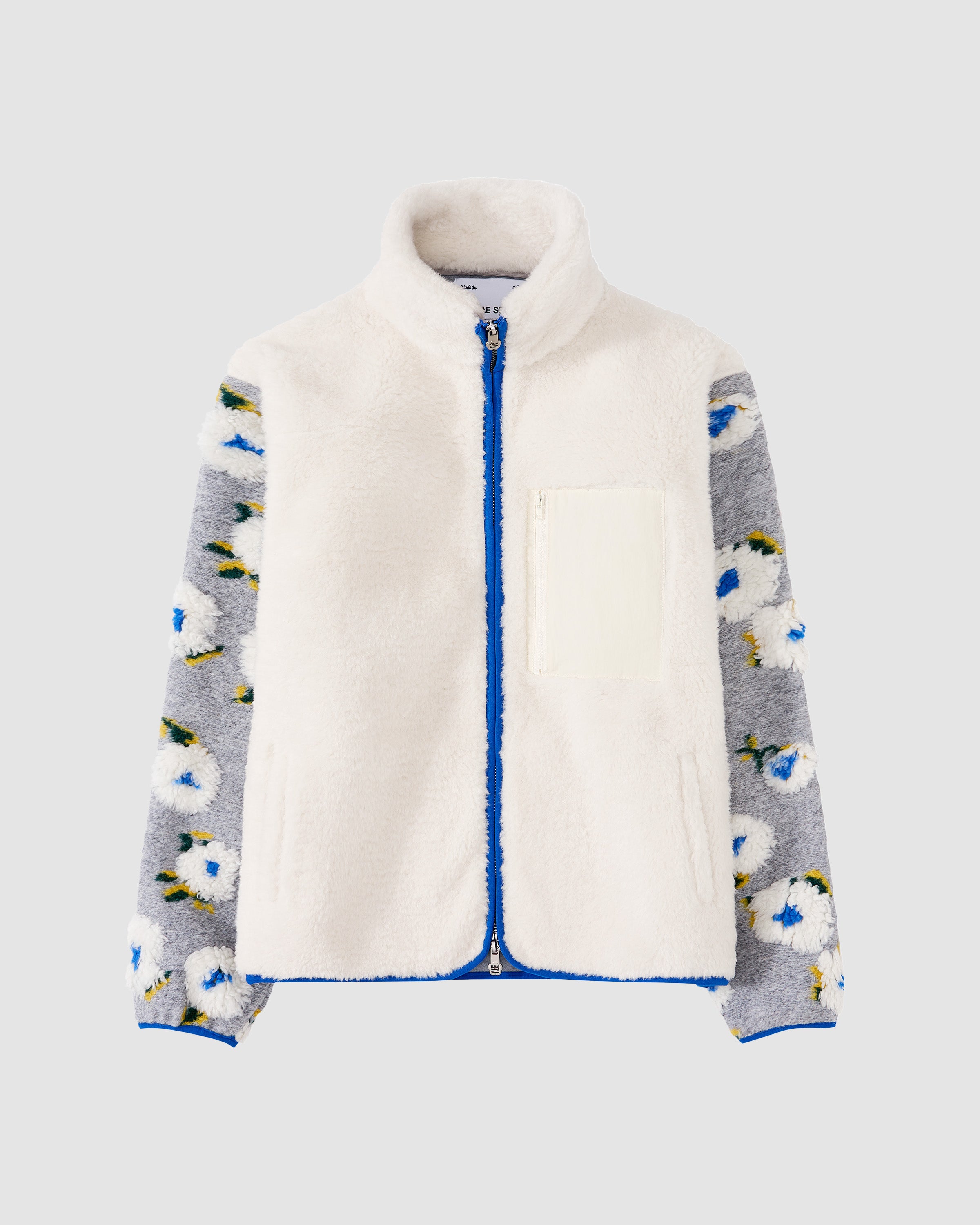 The IVORY MUGUNGHWA FLEECE by Sundae School features a white sherpa front with a high collar, grey hibiscus sleeves adorned with white and blue floral patterns, blue zipper, and two front pockets.