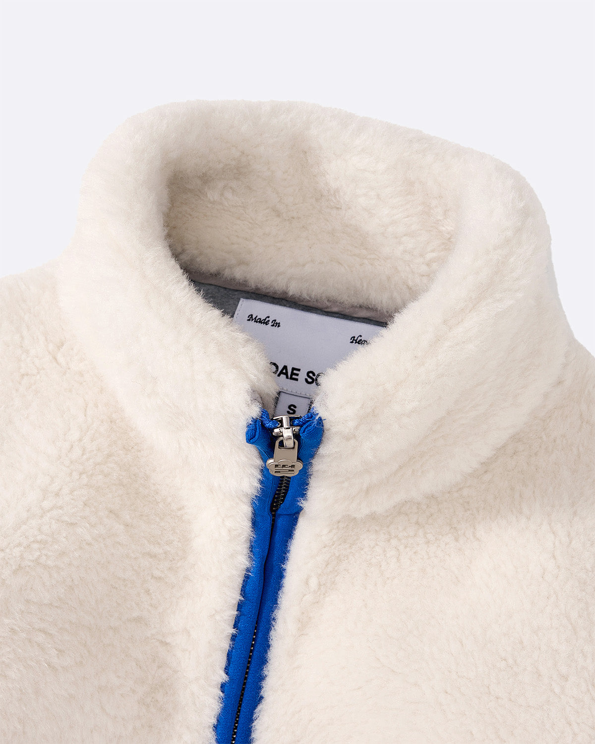 A close-up of the Sundae School IVORY MUGUNGHWA FLEECE, a white sherpa jacket with a high collar and blue zipper. The inner collar label is partly visible.