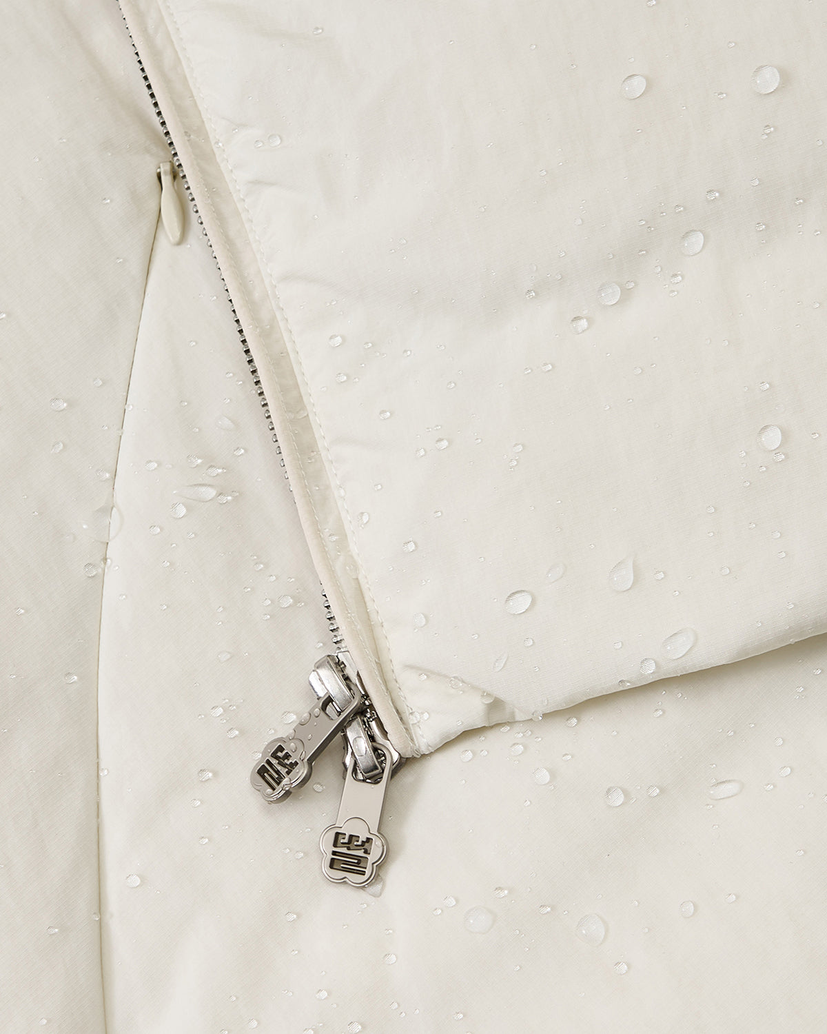 Close-up of the Sundae School IVORY LIME ORIGAMI PUFFER, a light-colored, water-resistant jacket featuring water droplets on the surface and a partially unzipped metal zipper.