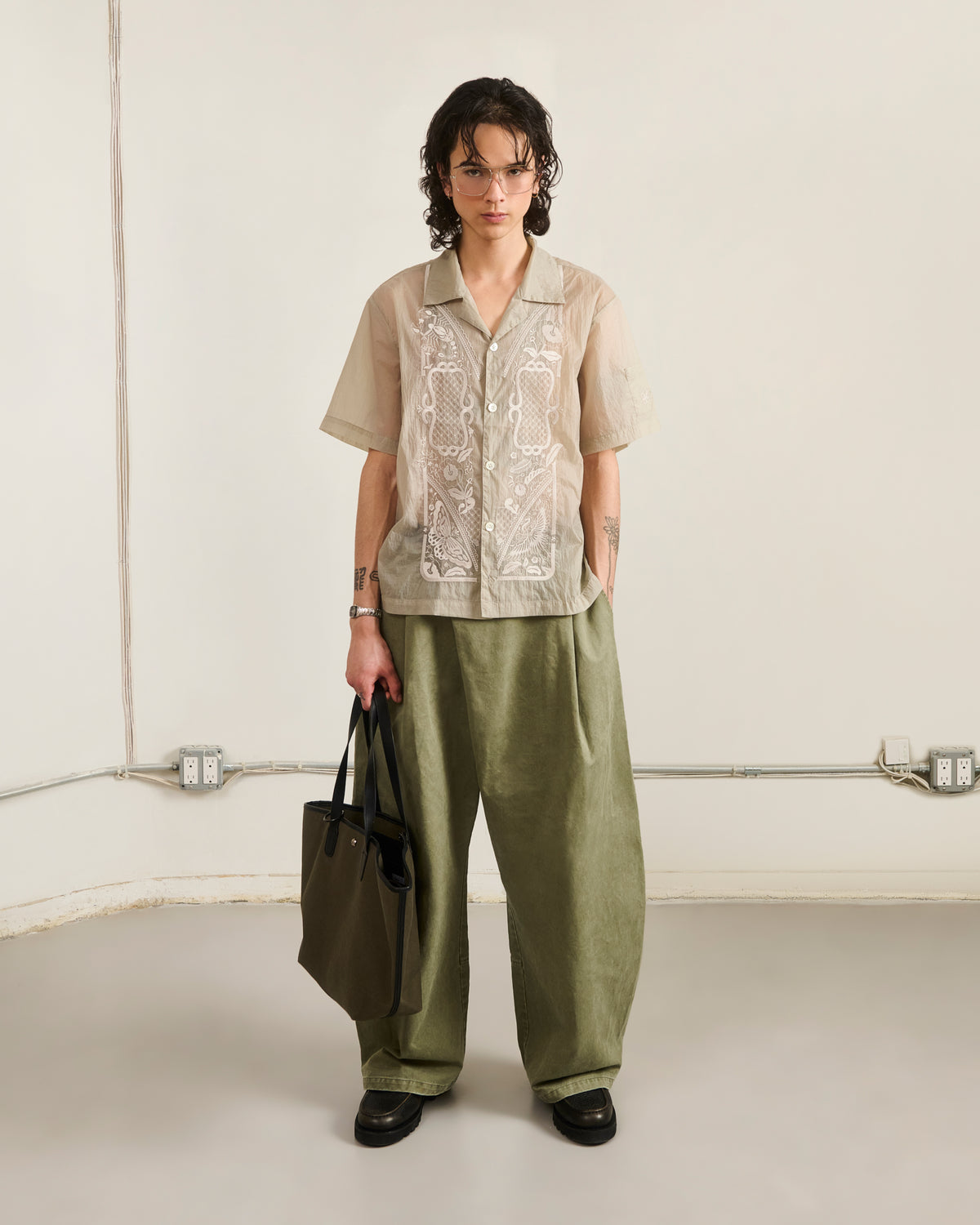 A person with short, wavy hair, glasses, and tattoos stands indoors against a neutral background. They wear a beige embroidered shirt and olive-green pants, holding Sundae School's OLIVE BOJAGI TIE TOTE BAG.