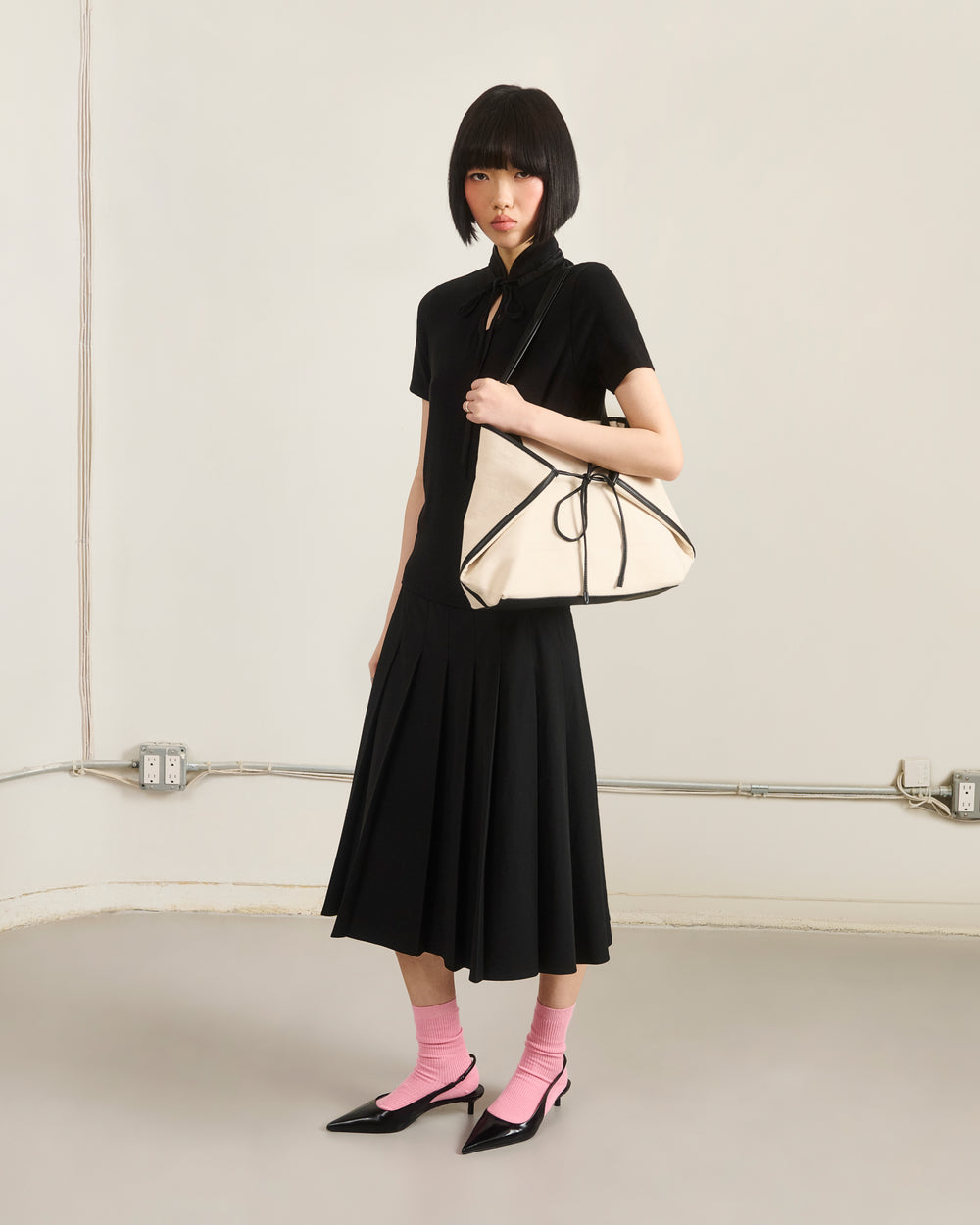 A woman with a short black bob wears a black dress, pink socks, and pointed black heels. She carries the Sundae School IVORY BOJAGI TIE TOTE BAG with vegan leather handles, standing against a plain wall with exposed outlets and pipes.