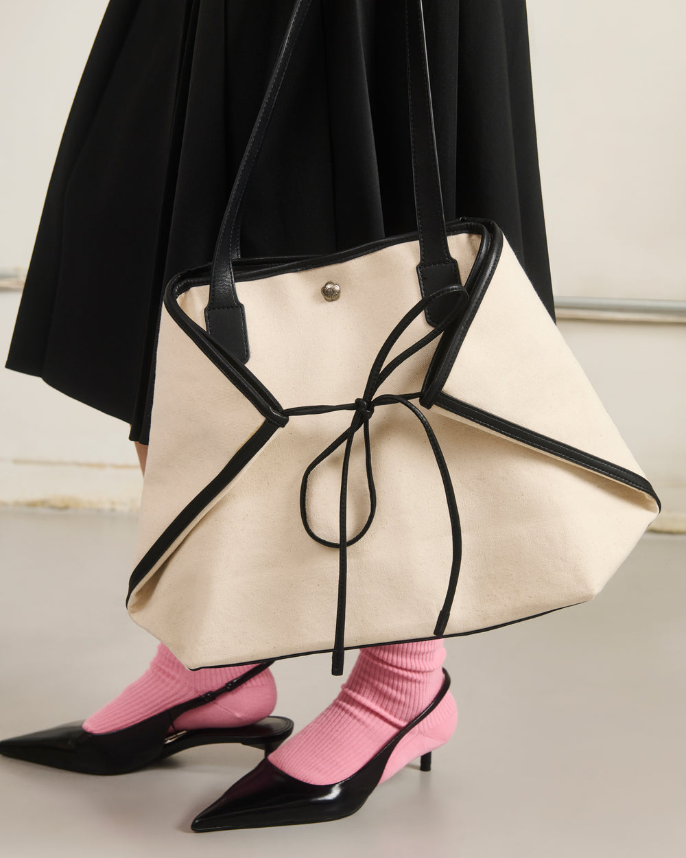 A person in a black skirt, pink socks, and black heels carries the Sundae School IVORY BOJAGI TIE TOTE BAG, featuring black trim, vegan leather handles, and a decorative black tie.