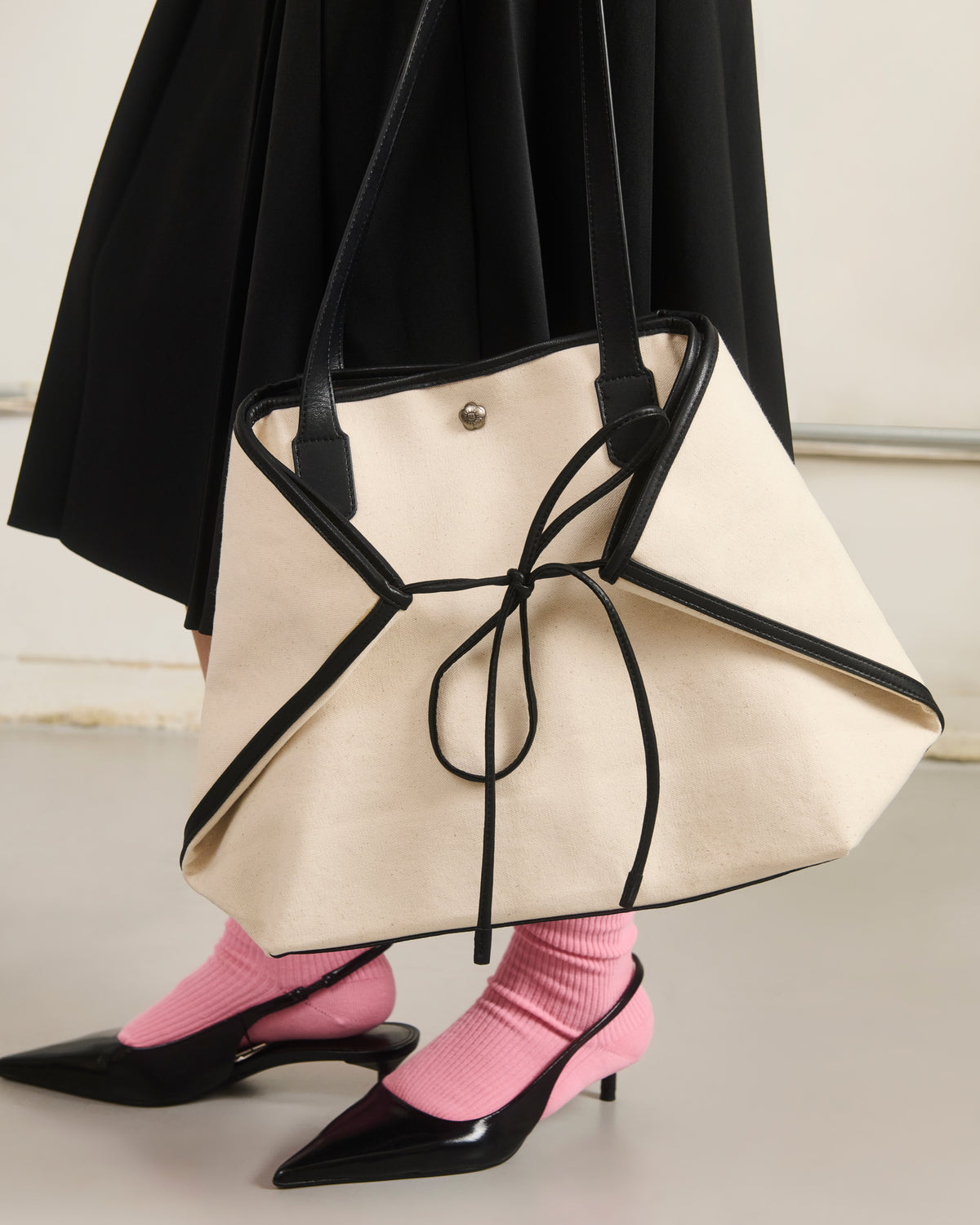 A person in a black skirt, pink socks, and black heels carries the Sundae School IVORY BOJAGI TIE TOTE BAG, featuring black trim, vegan leather handles, and a decorative black tie.