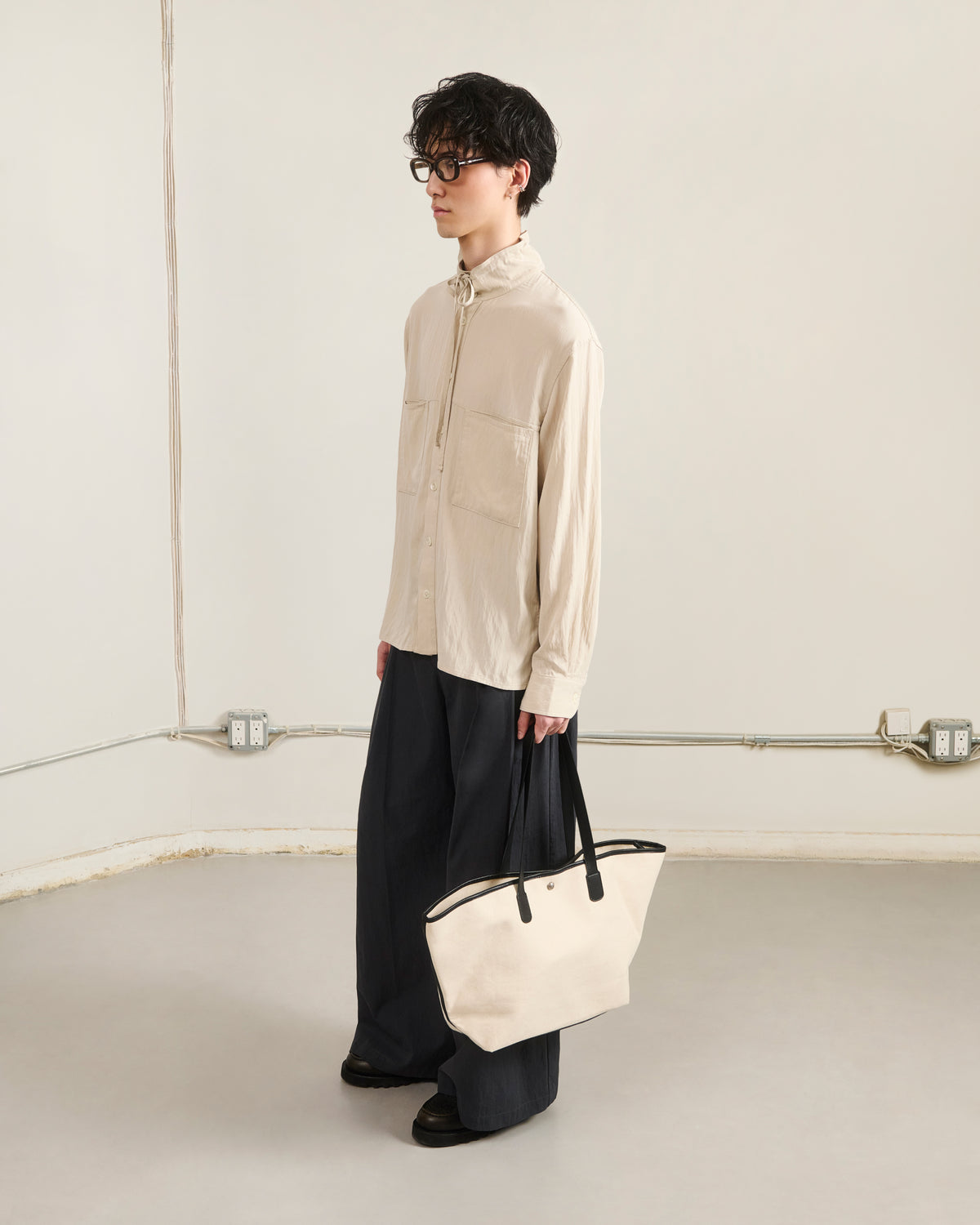 A person with short dark hair, glasses, and a neutral expression stands indoors wearing a beige zip-up shirt, dark pants, black shoes, and holding the Sundae School IVORY BOJAGI TIE TOTE BAG with vegan leather handles.