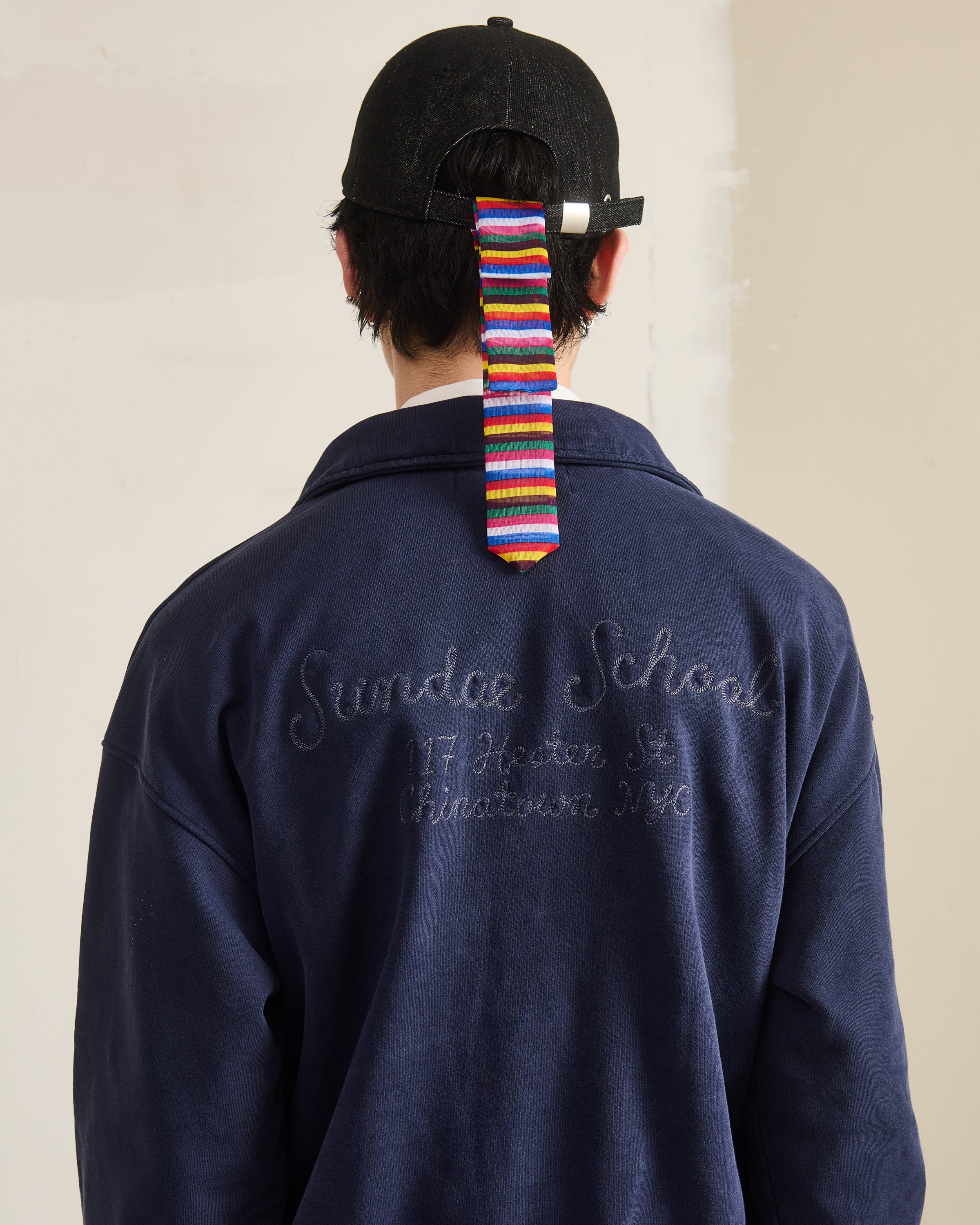 A person in a dark baseball cap with a colorful striped ribbon, seen from behind, wears the Sundae School NAVY HESTER QUARTER ZIP with “Sundae School, 117 Hester St, Chinatown NYC” embroidered on the back.