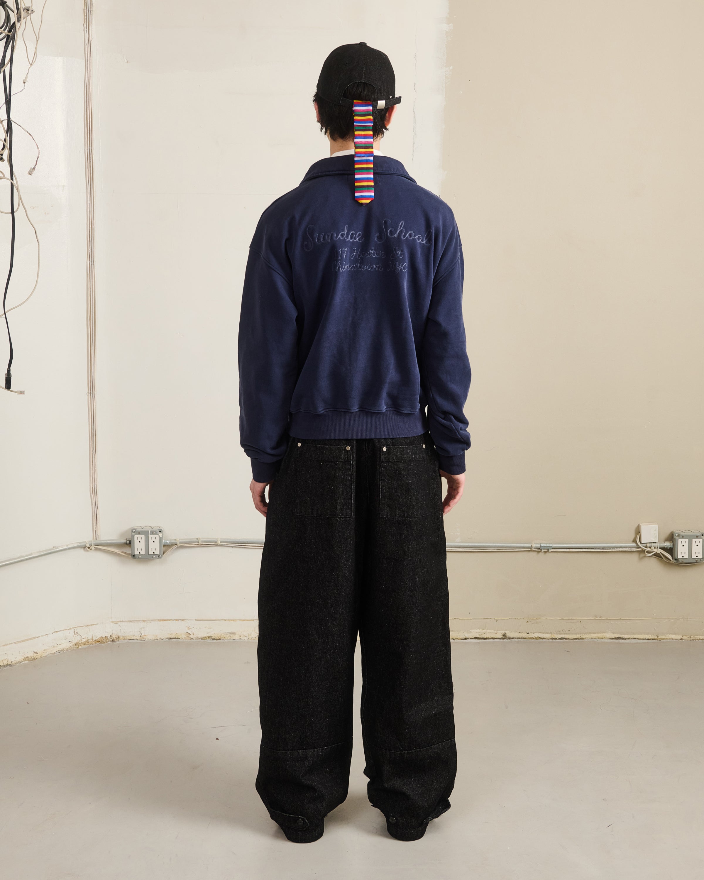 A person faces a beige wall in the Sundae School NAVY HESTER QUARTER ZIP, wide black pants, and a black cap with a colorful striped strap. Exposed wiring and electrical outlets appear on the wall behind them.