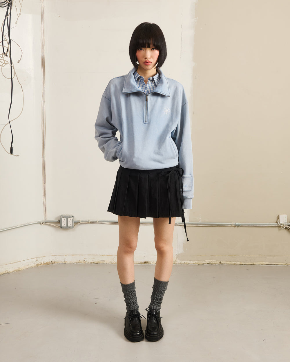 A person with a bob stands indoors against a plain wall, wearing the Sundae School SKY BLUE HESTER QUARTER ZIP over a blue checkered shirt, paired with a black pleated skirt, gray socks, and black shoes.